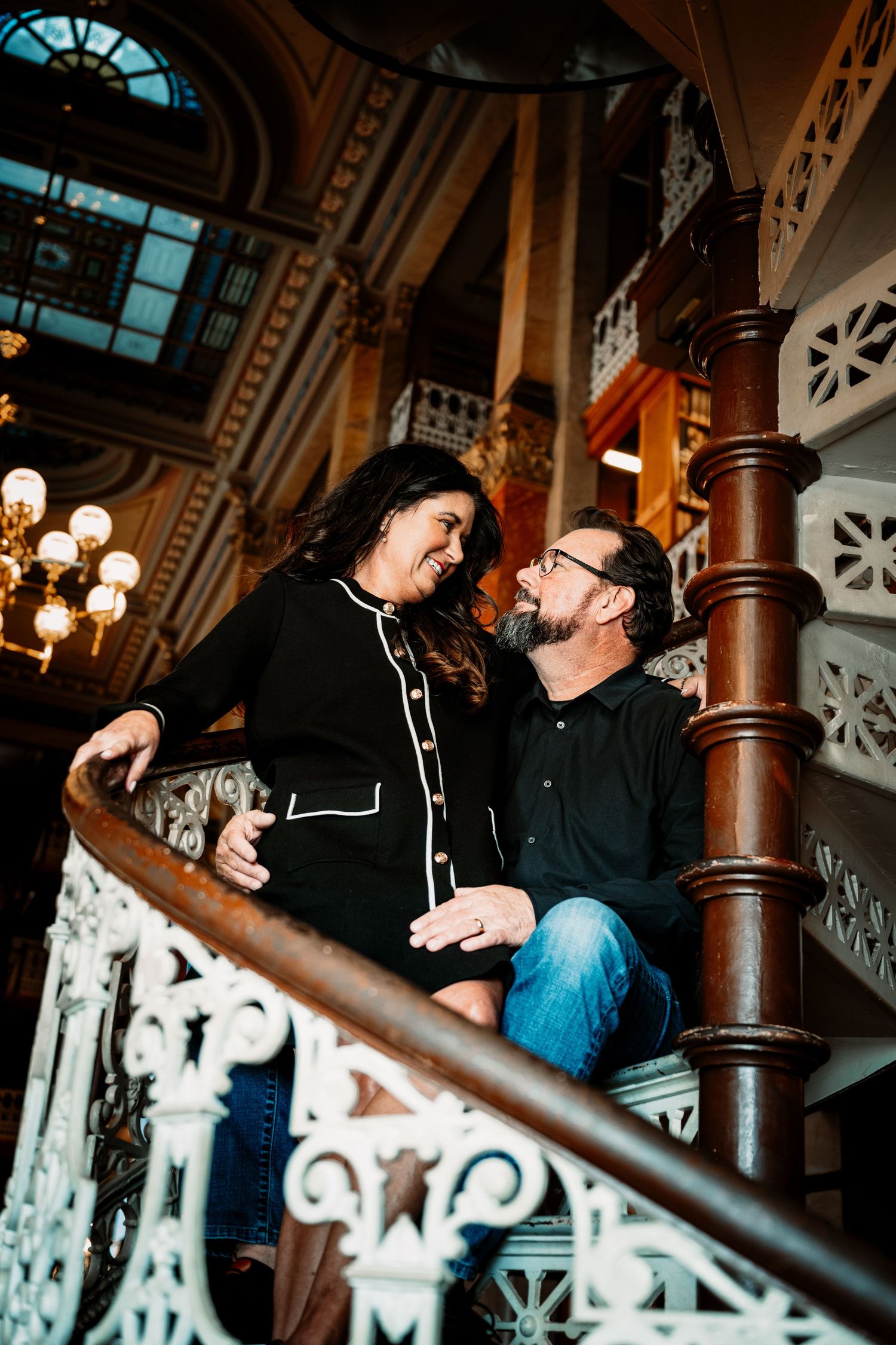 Couple laughing together on ornate white wrought iron staircase with wooden banister in elegant historic building.