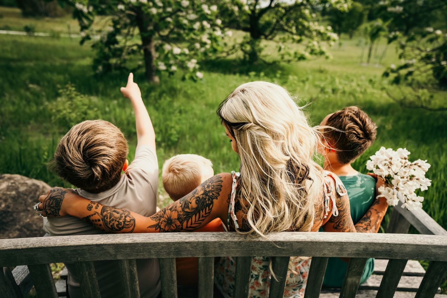 Family sitting on a wooden bench in a lush green park, with tattooed mom and three children enjoying nature.