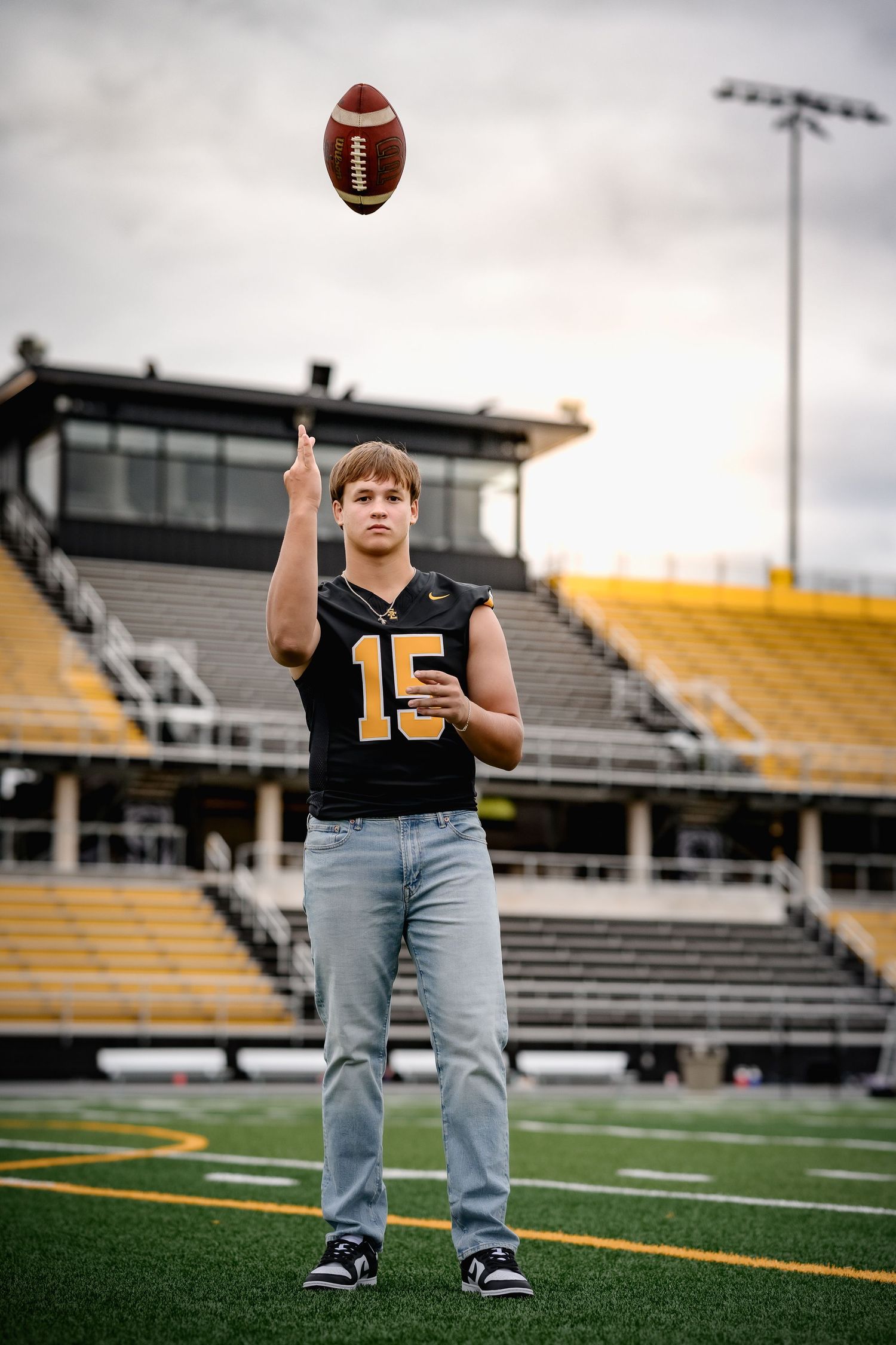 High school football player in black number 15 jersey tosses a ball on stadium field under cloudy sky.