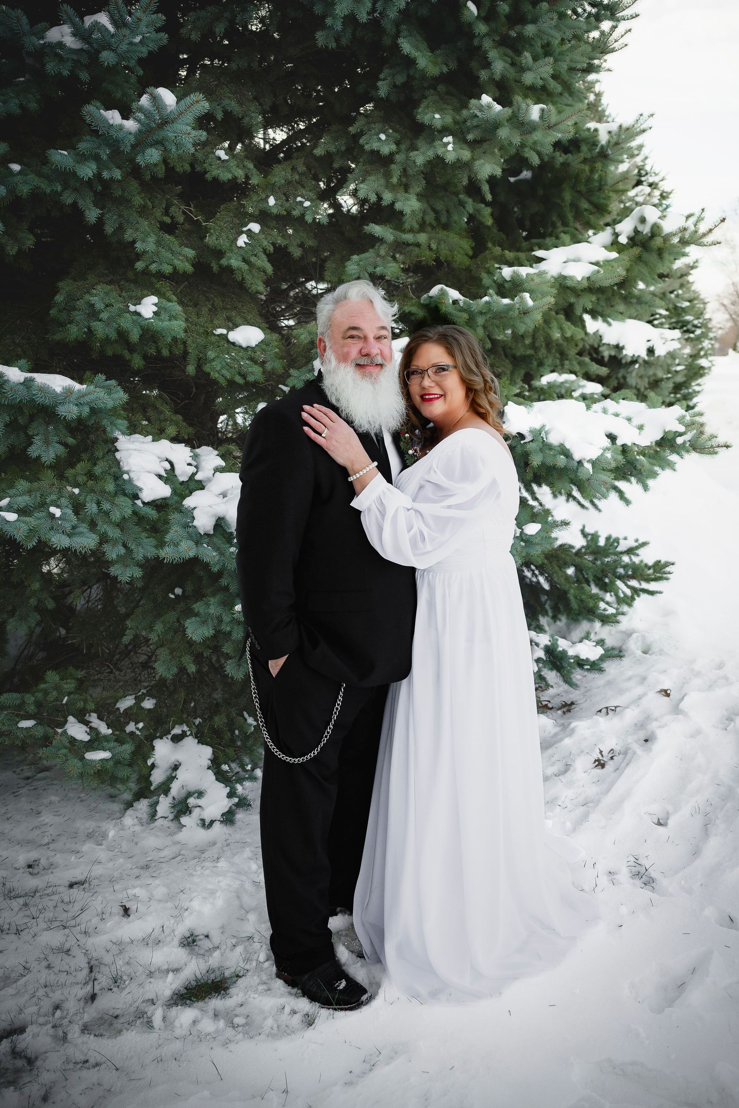 Older couple in wedding attire embracing in front of a snow-covered evergreen tree outdoors.