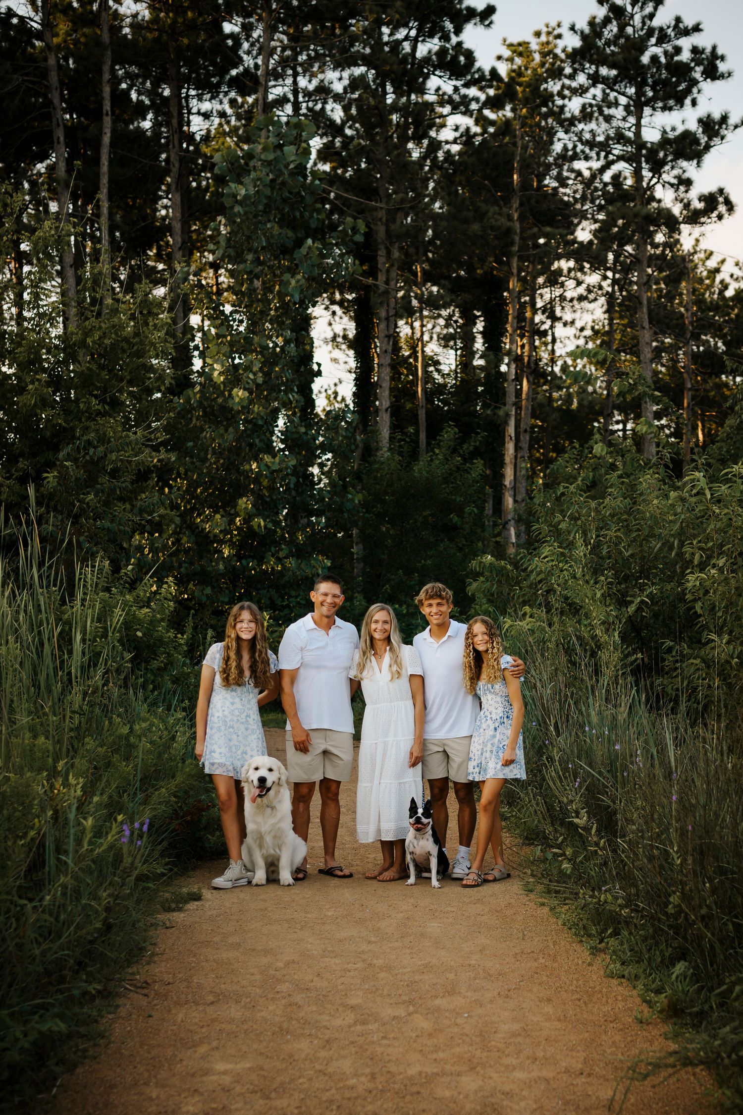 Family of five dressed in white posing with two dogs on a wooded trail at golden hour.