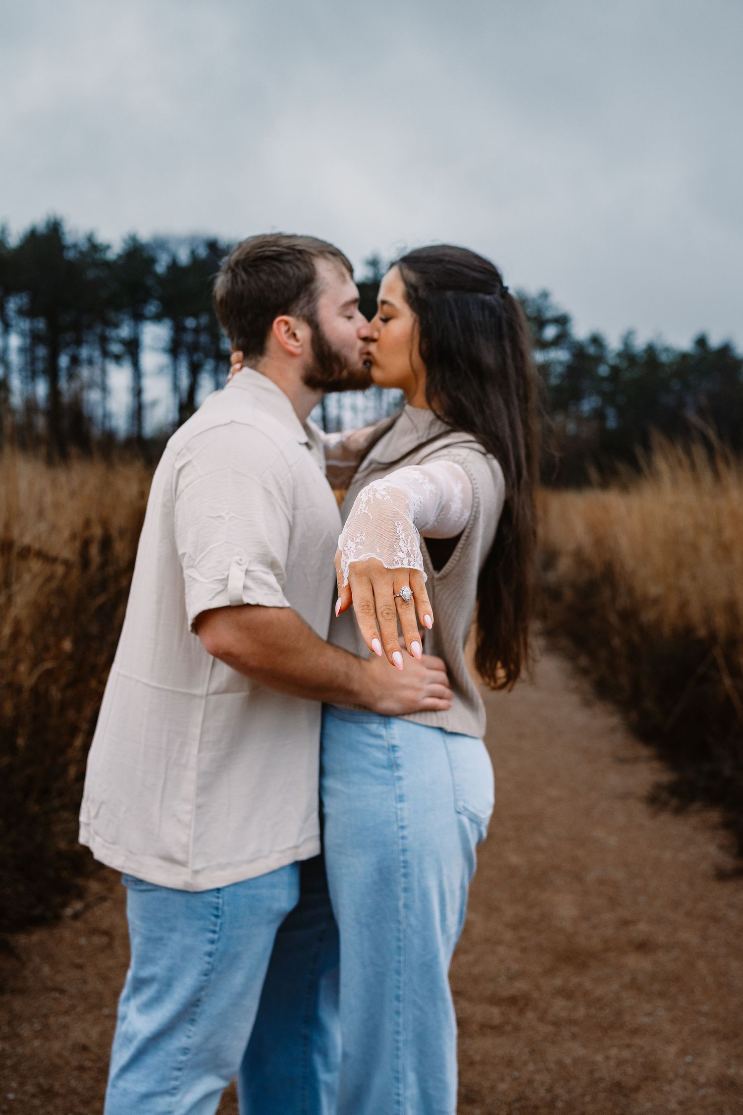 Engaged couple kissing on outdoor path, woman displaying diamond engagement ring to camera.