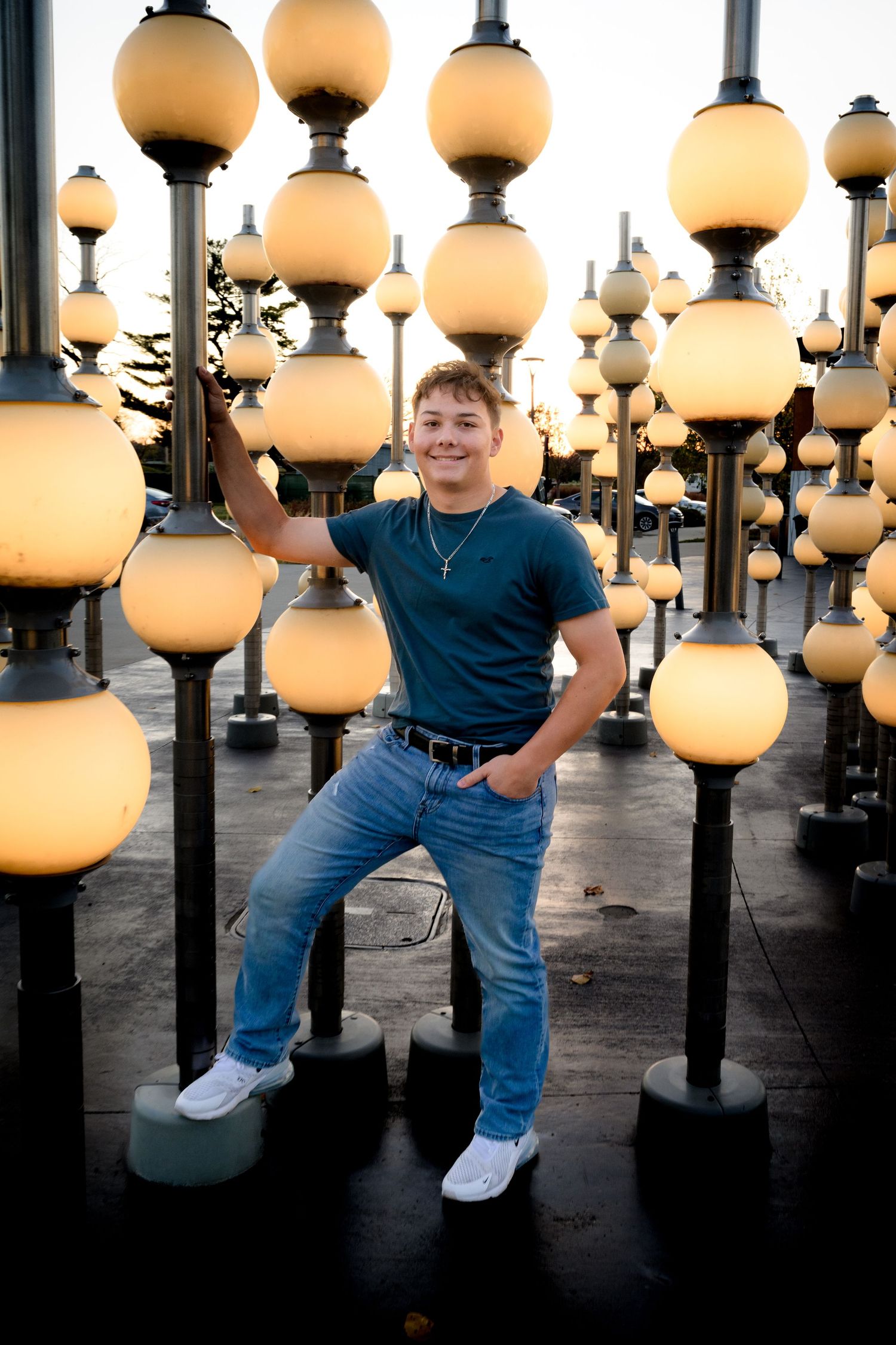 Teenage boy posing confidently among glowing globe street lamps at dusk outdoors.
