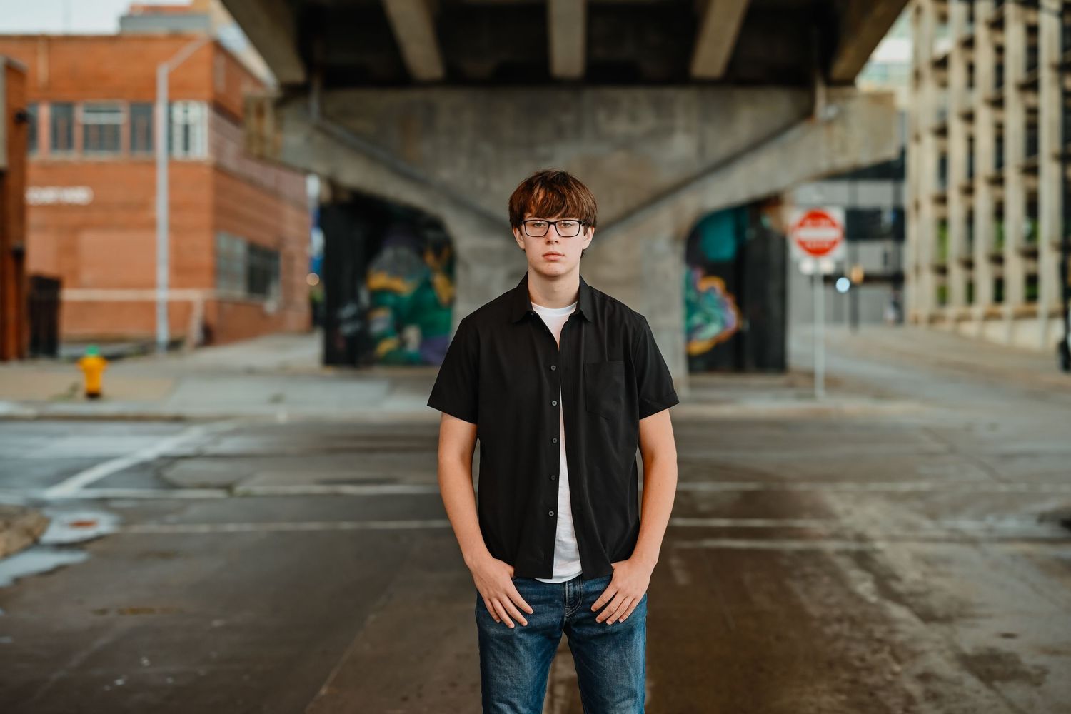 Young man in black shirt standing under urban bridge with colorful graffiti murals.