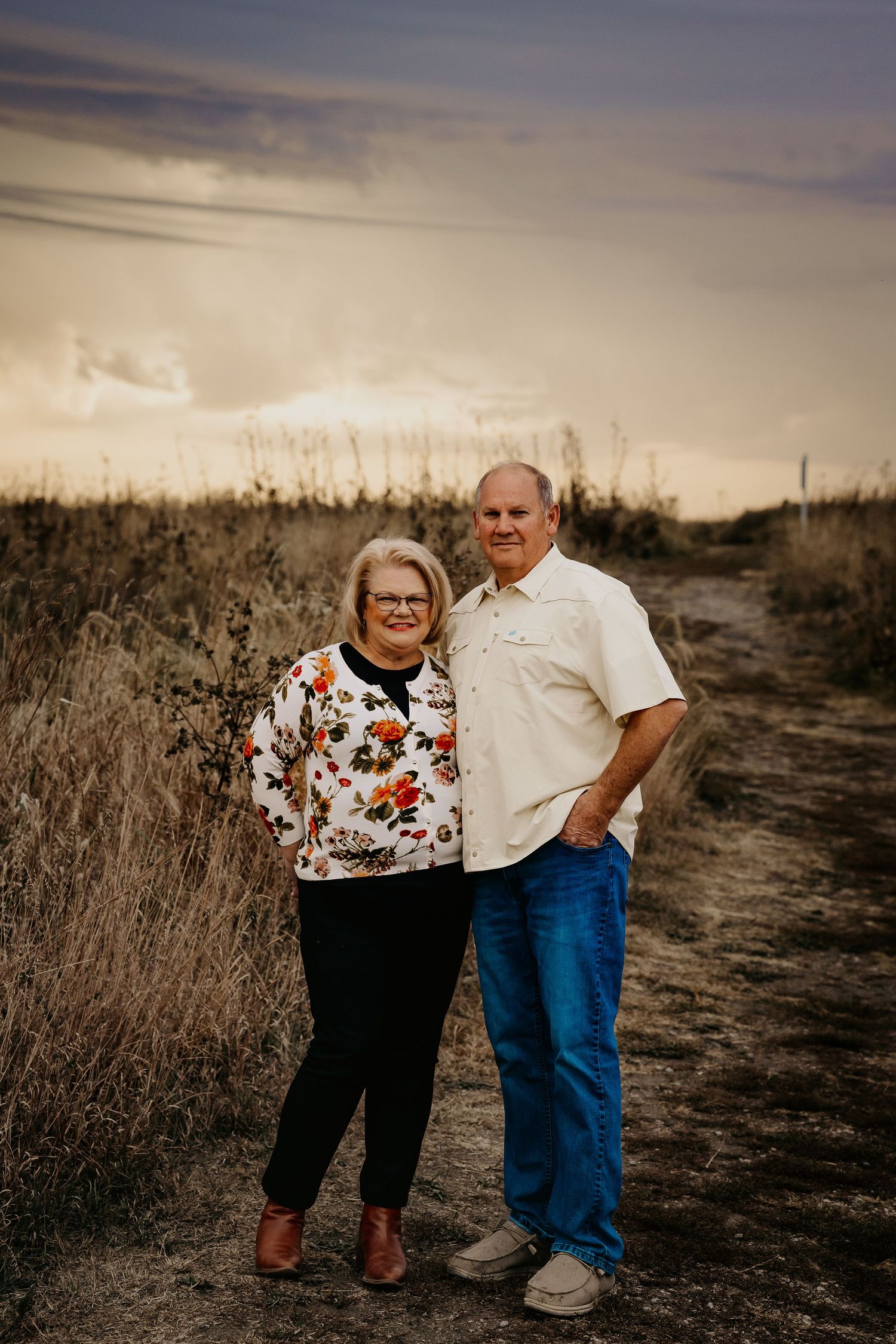 Older couple smiling together on a dirt path surrounded by golden fields at sunset.
