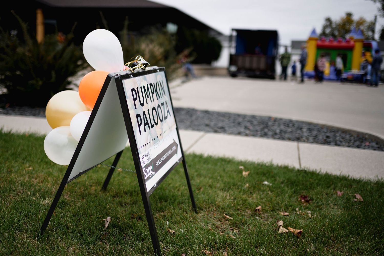 A Pumpkin Palooza event sign with orange and white balloons outside a community gathering with a bounce house.