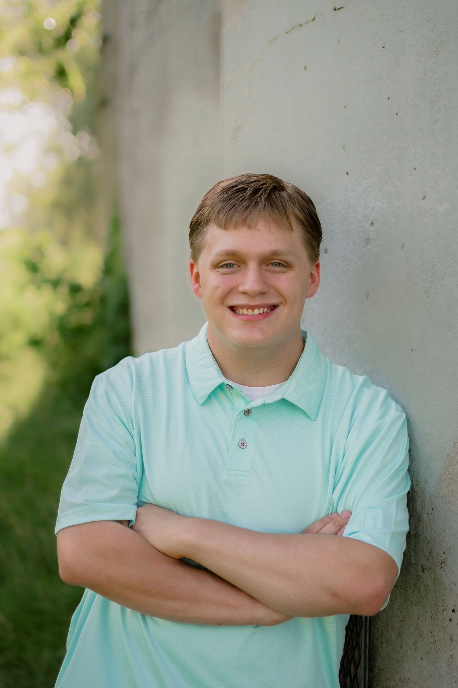 Smiling young man in mint polo shirt leans against concrete wall with arms crossed outdoors.