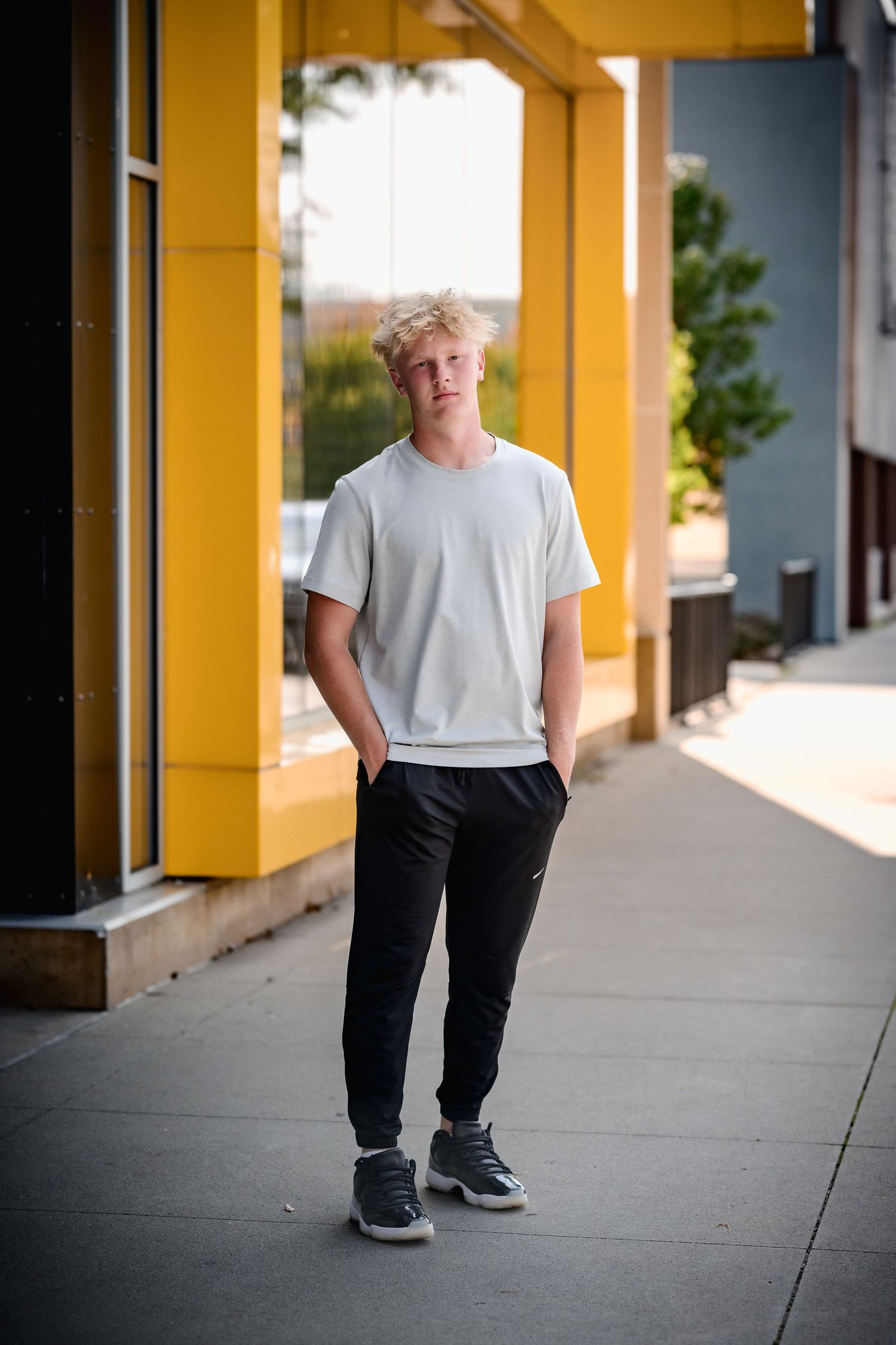 Blond teenage boy in white t-shirt and black joggers standing on urban sidewalk.
