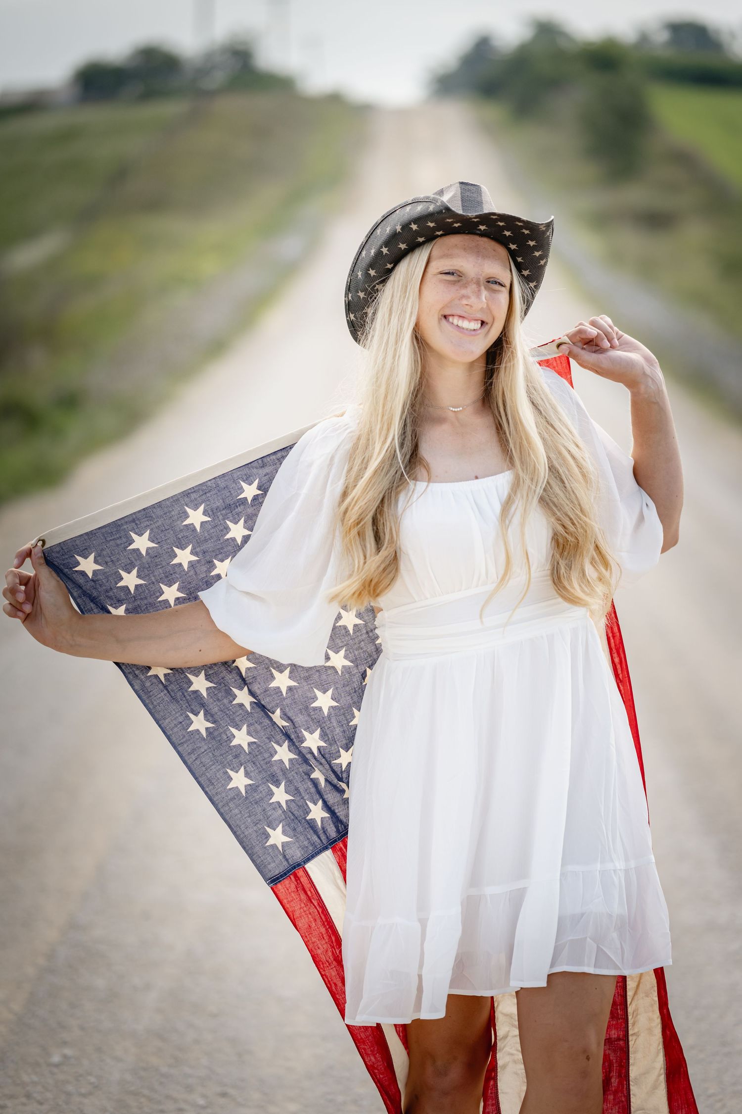 Smiling blonde woman in white dress and cowboy hat holding American flag on rural dirt road.