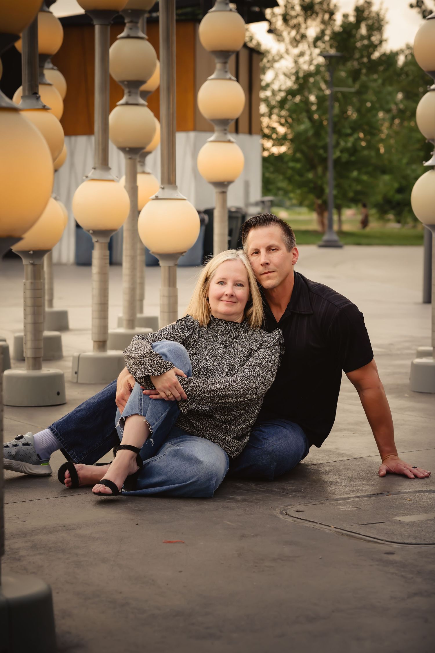 Couple sitting on ground surrounded by globe lights at dusk, woman leaning against man in casual jeans and tops.