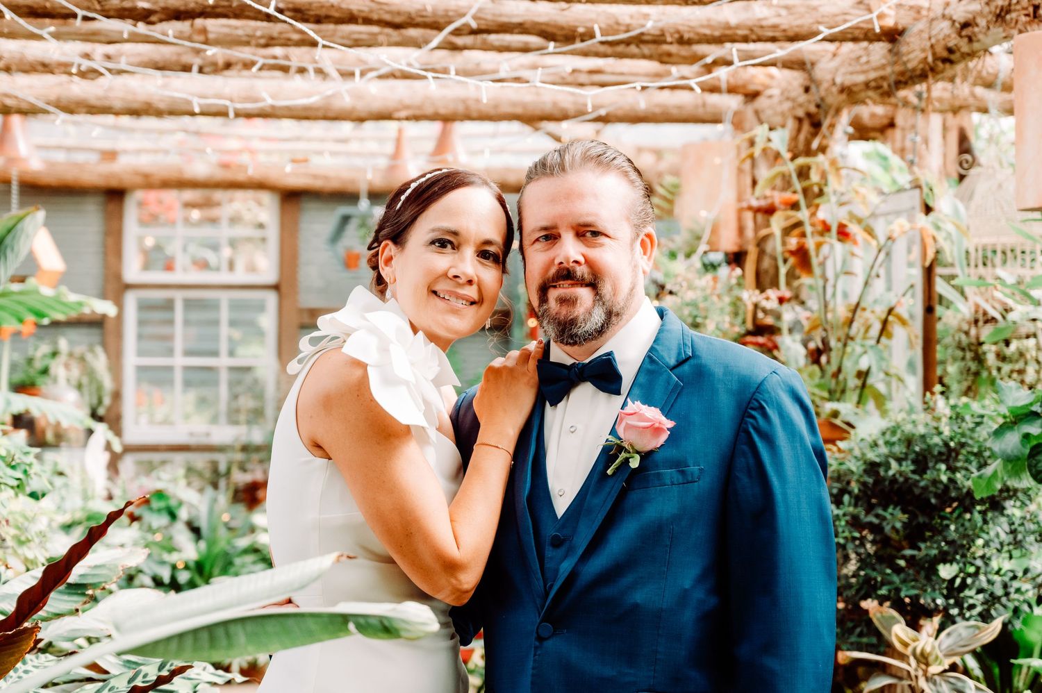 Smiling bride and groom pose together in botanical garden venue with lush greenery backdrop.