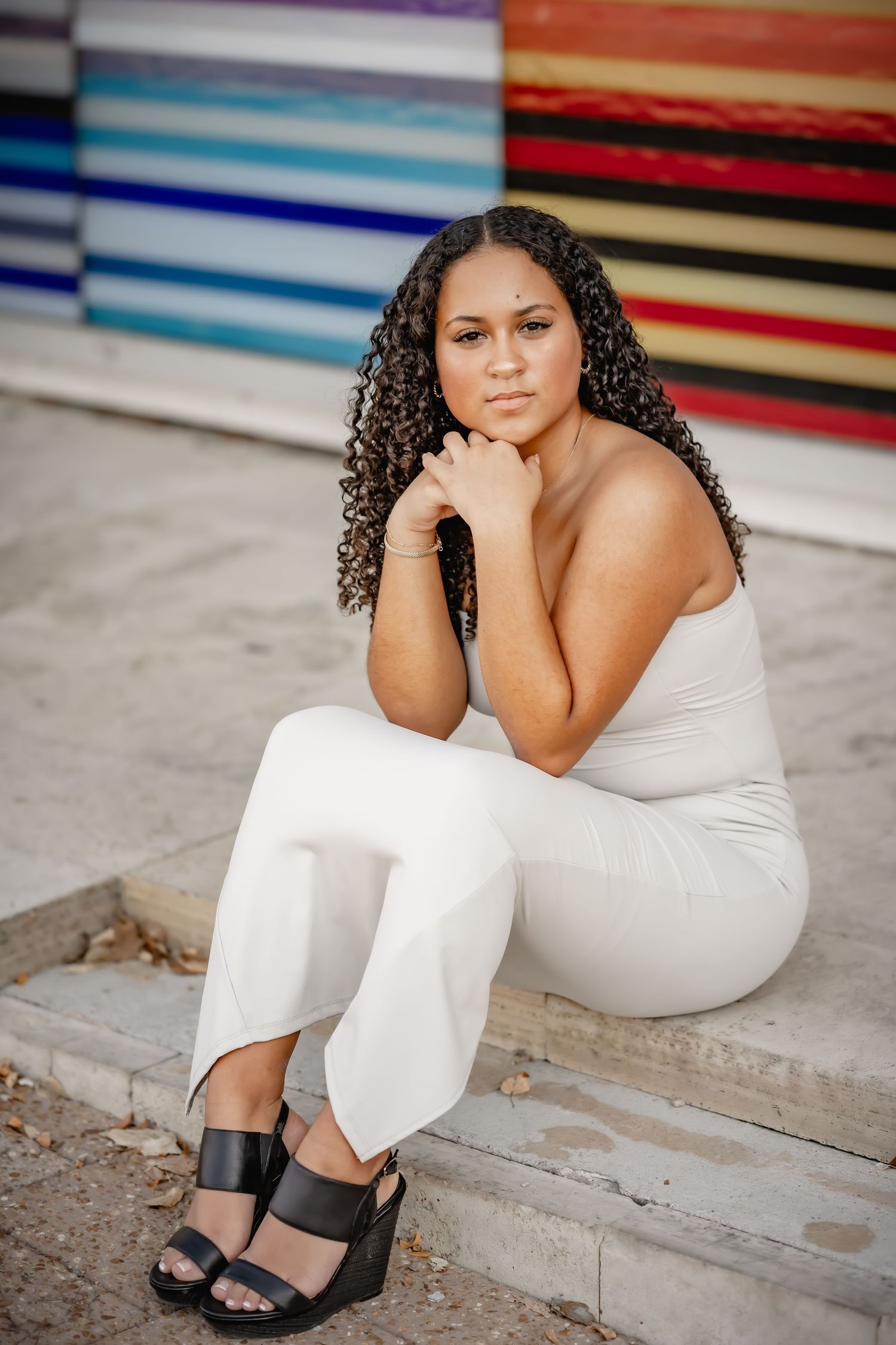Young woman with curly hair in white outfit sitting on steps before colorful striped wall.