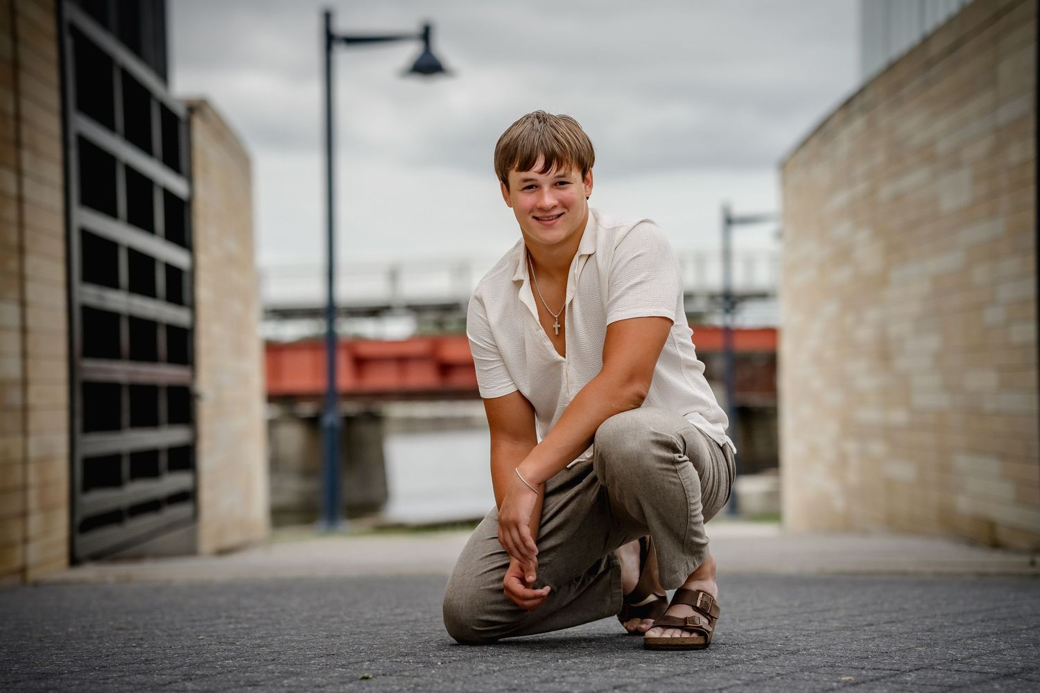 Person crouching outdoors on brick pathway between modern buildings, wearing white shirt and khaki pants.