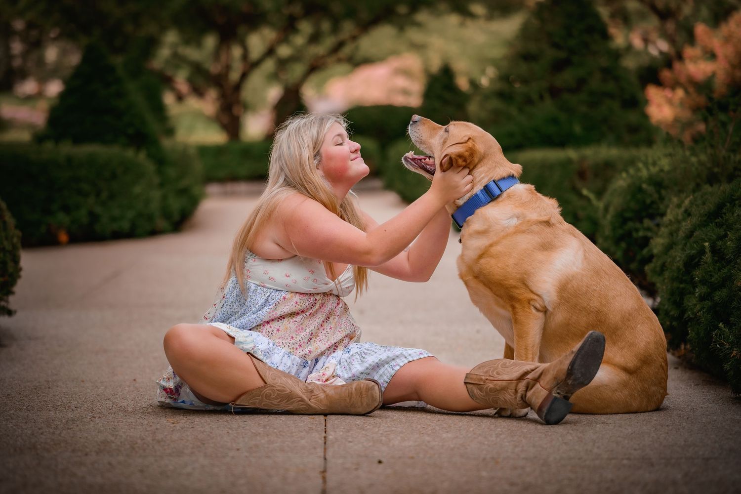 Young blonde woman laughing joyfully while playing with a golden Labrador dog on an outdoor pathway.