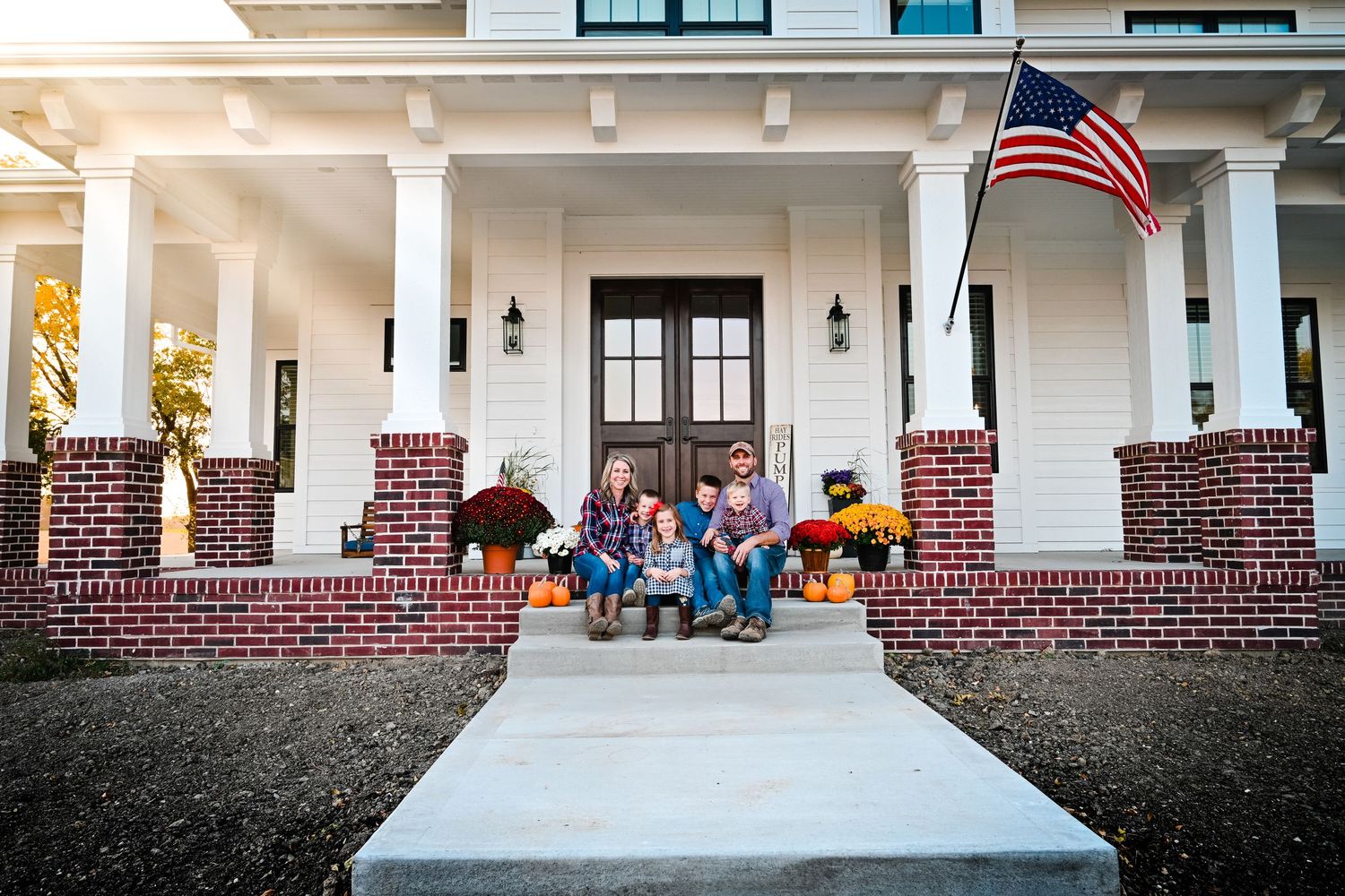 Family of three sitting on front porch steps of white farmhouse decorated with fall pumpkins and American flag.