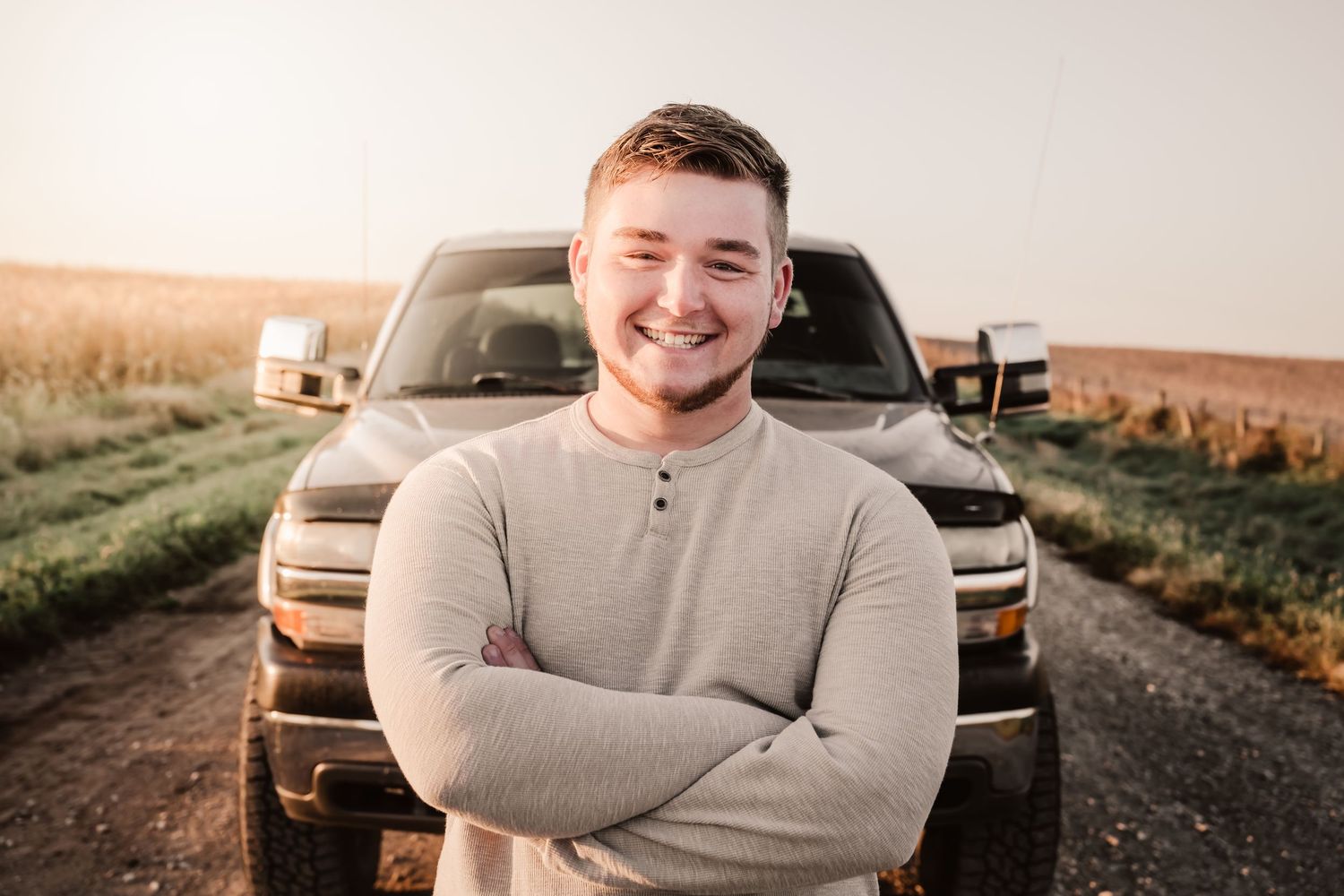 Smiling young man with arms crossed standing in front of a truck at sunset.