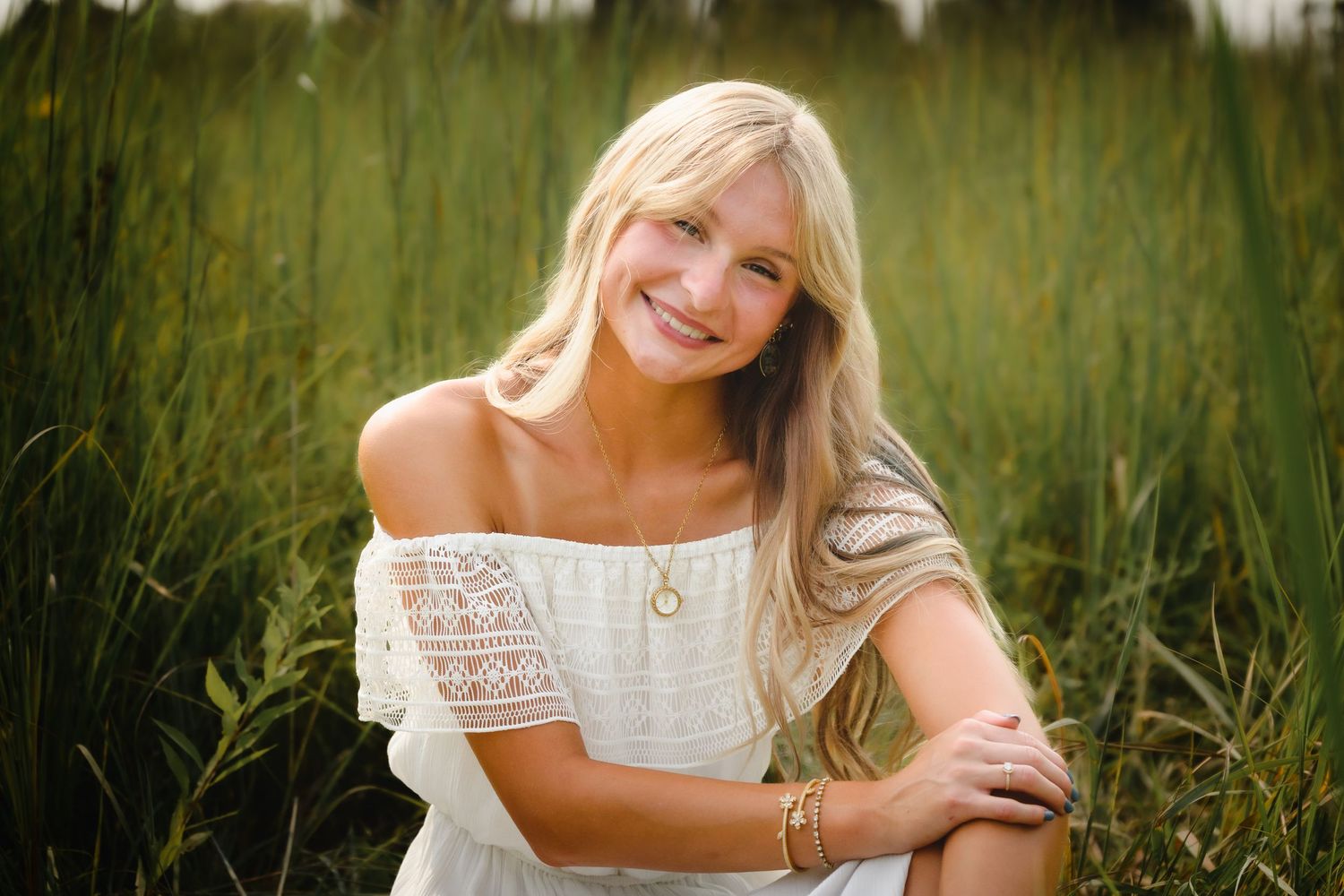 Smiling blonde woman in white off-shoulder top sitting in lush green field during golden hour.