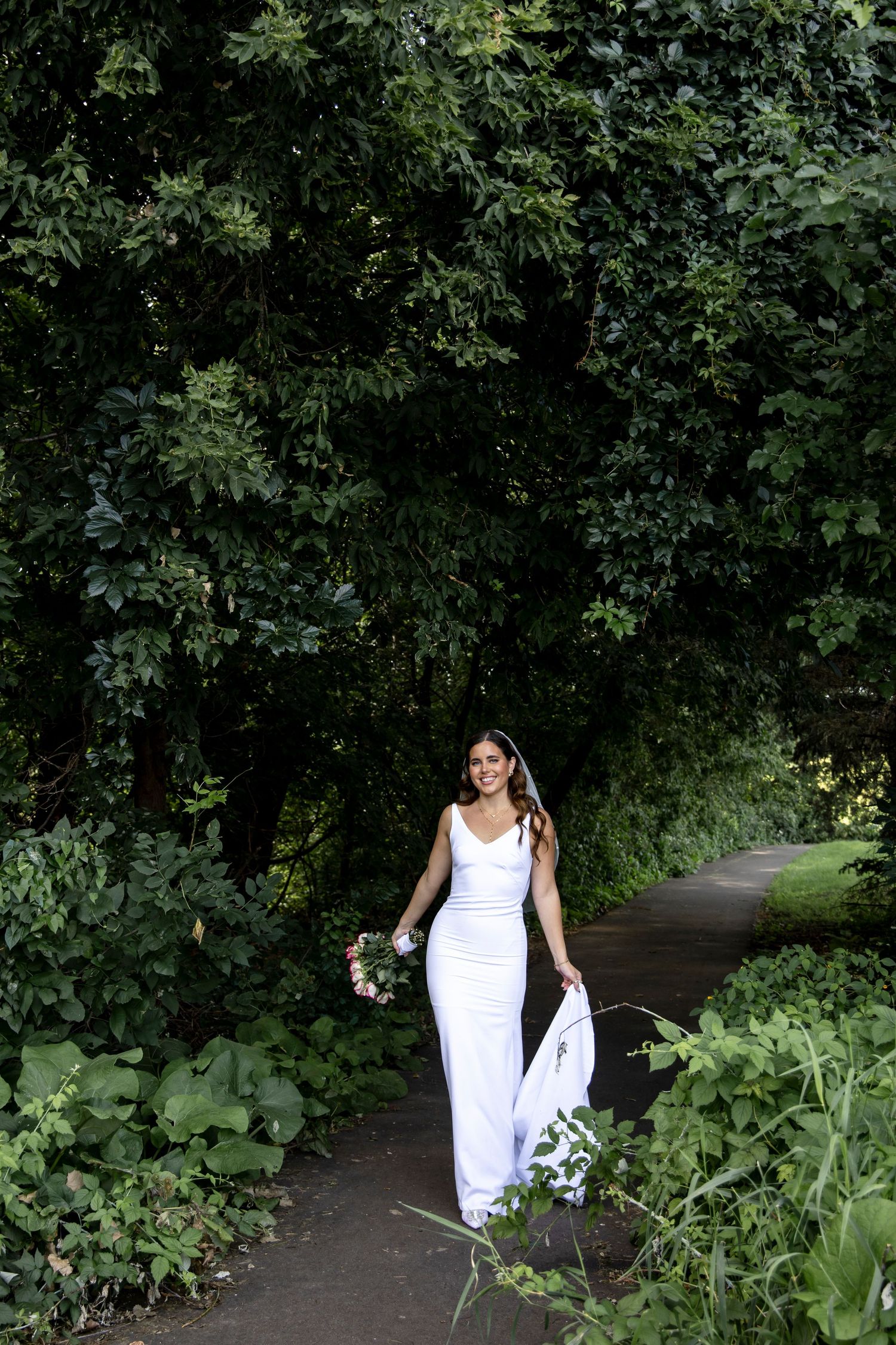 A person in a white dress walks along a garden path surrounded by lush greenery.