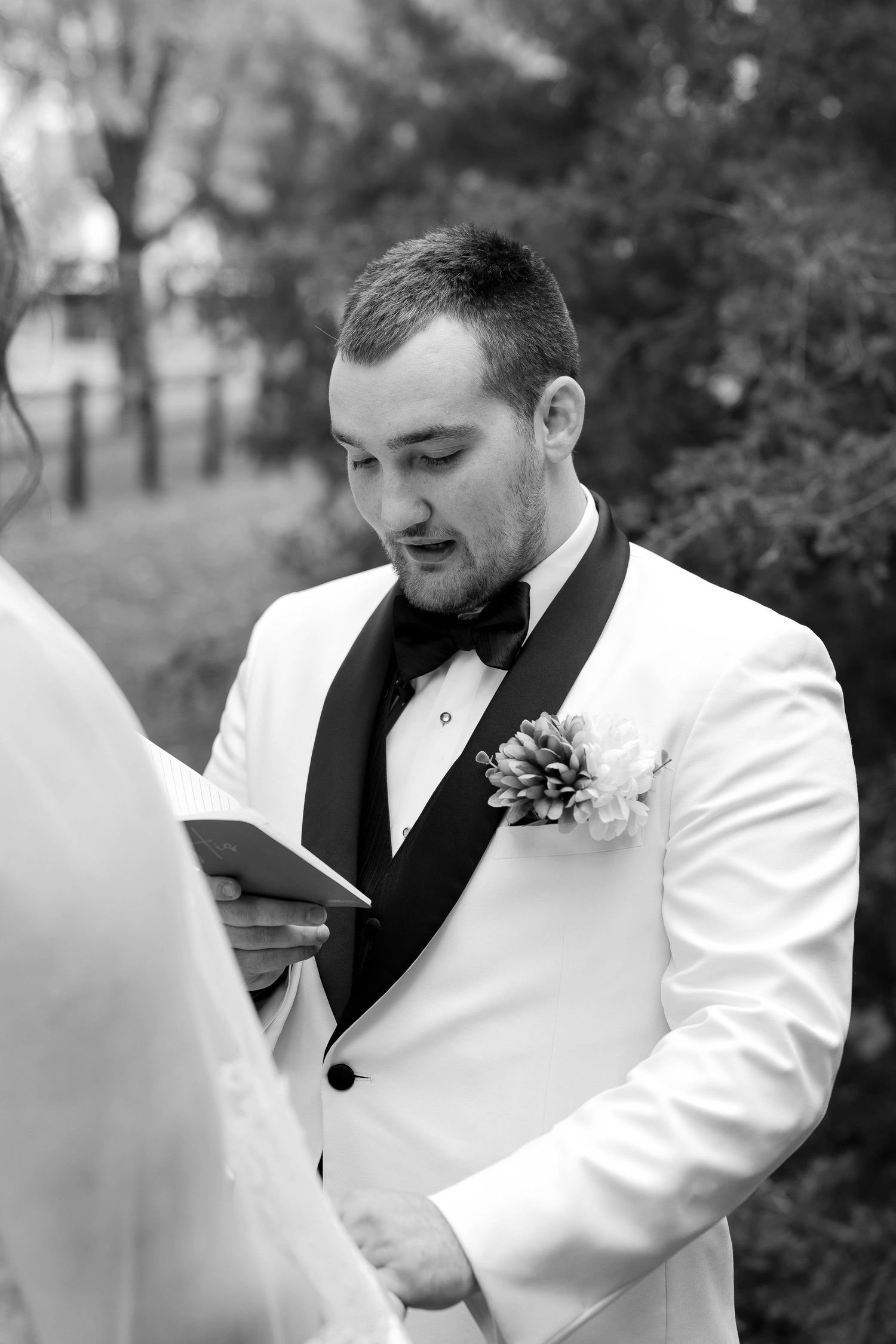 Groom in white tuxedo jacket reading during outdoor ceremony.
