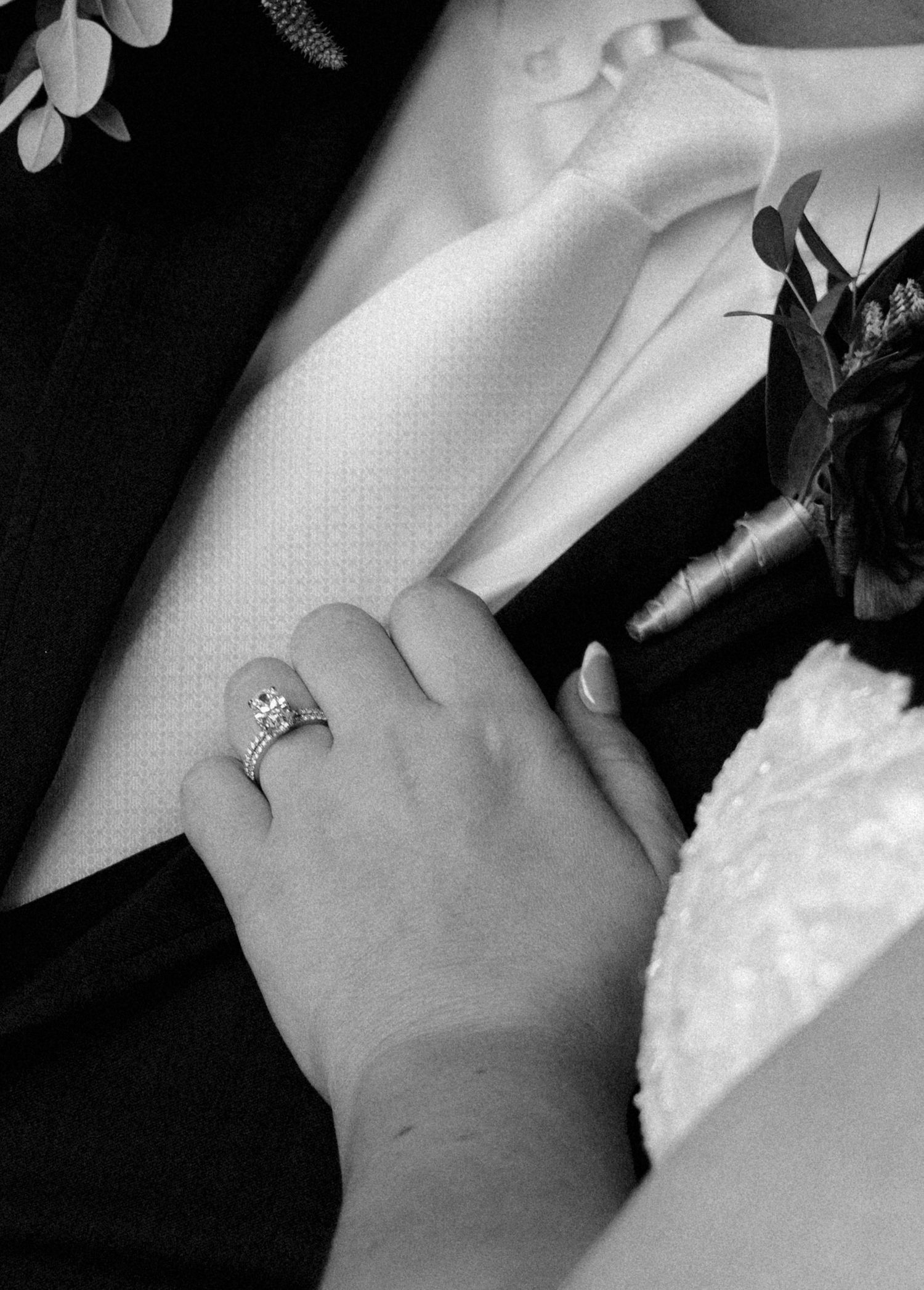 Black and white wedding photo of bride's hand with diamond engagement ring resting on groom's suit lapel with boutonniere.
