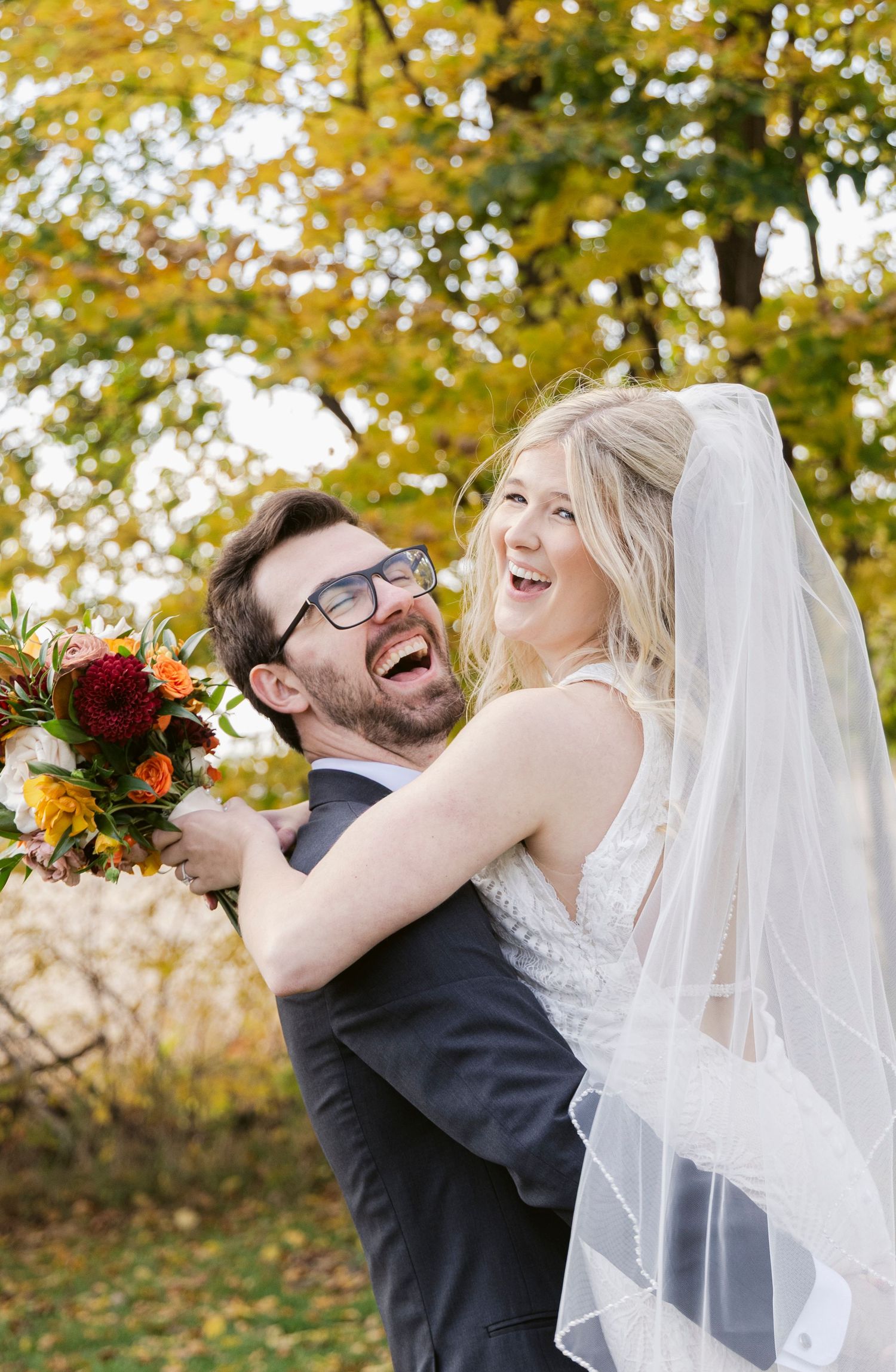 A wedding couple shares laughter and embraces against a backdrop of autumn trees.