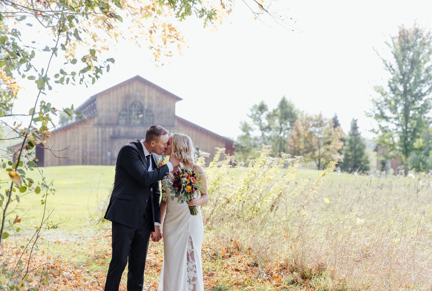 Wedding couple walking together in a scenic autumn field with barn in background.