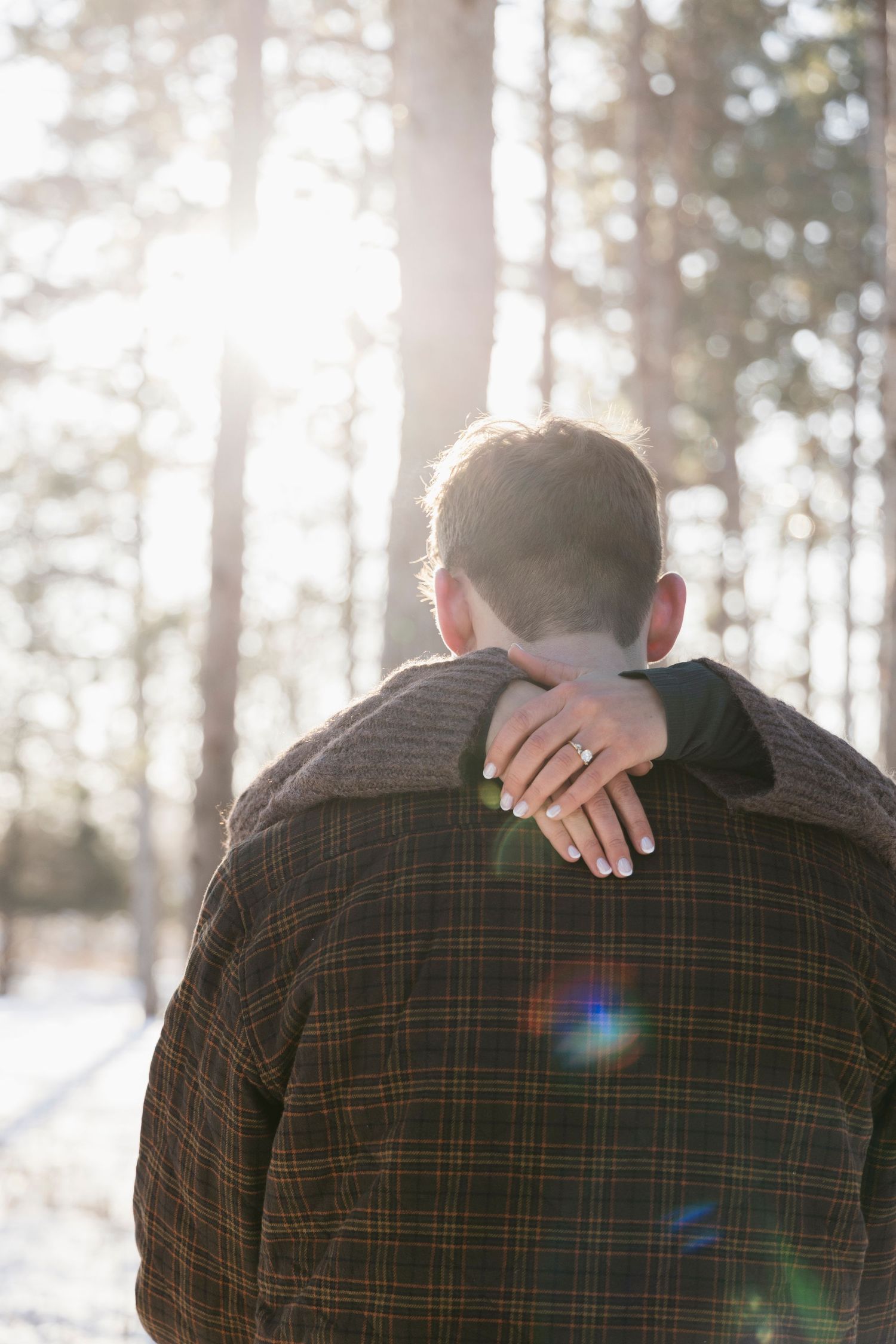 Back view of couple in plaid shirt embracing in sunlit winter forest.