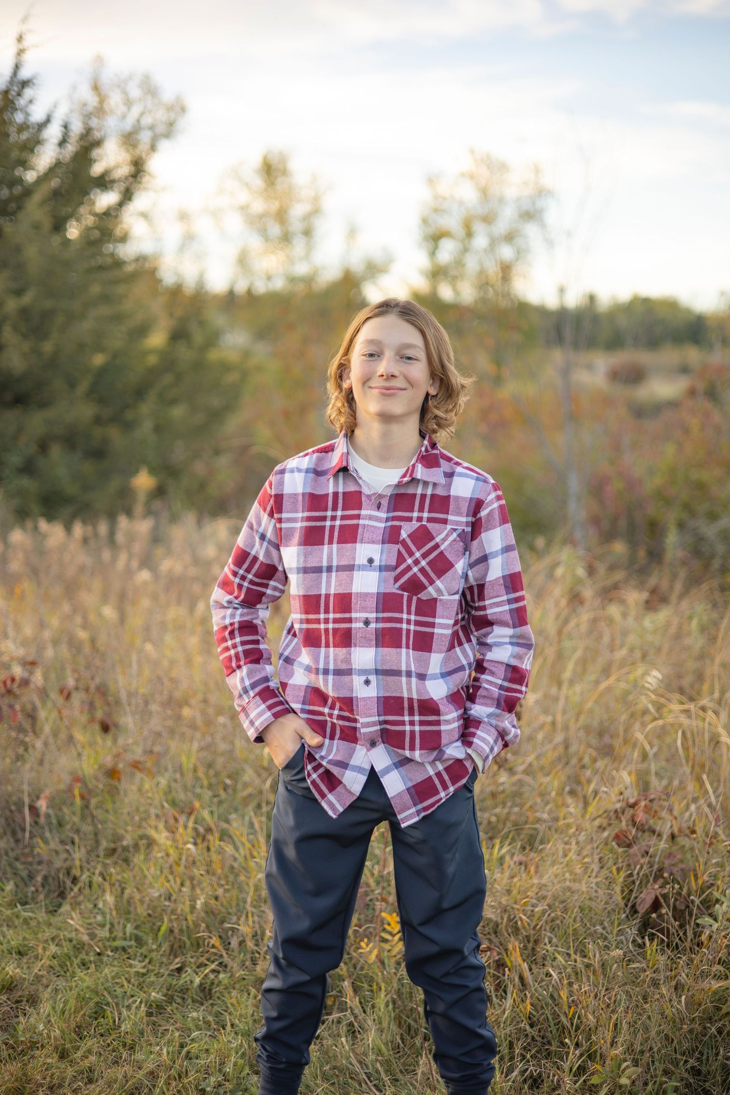 Person in red plaid shirt and jeans stands confidently in tall grass during outdoor photoshoot.