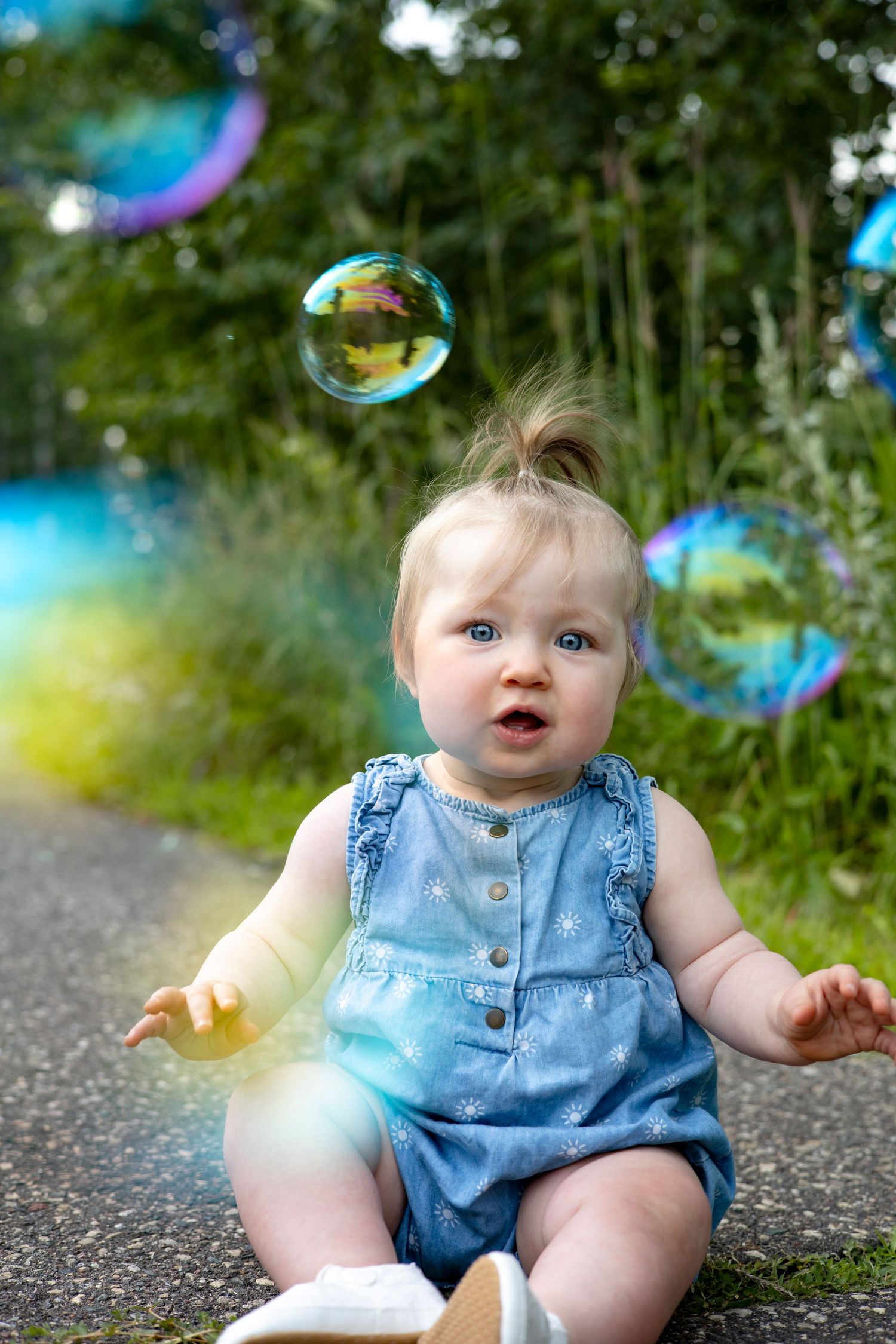 Baby in denim romper plays with colorful soap bubbles on a sunny day outdoors.