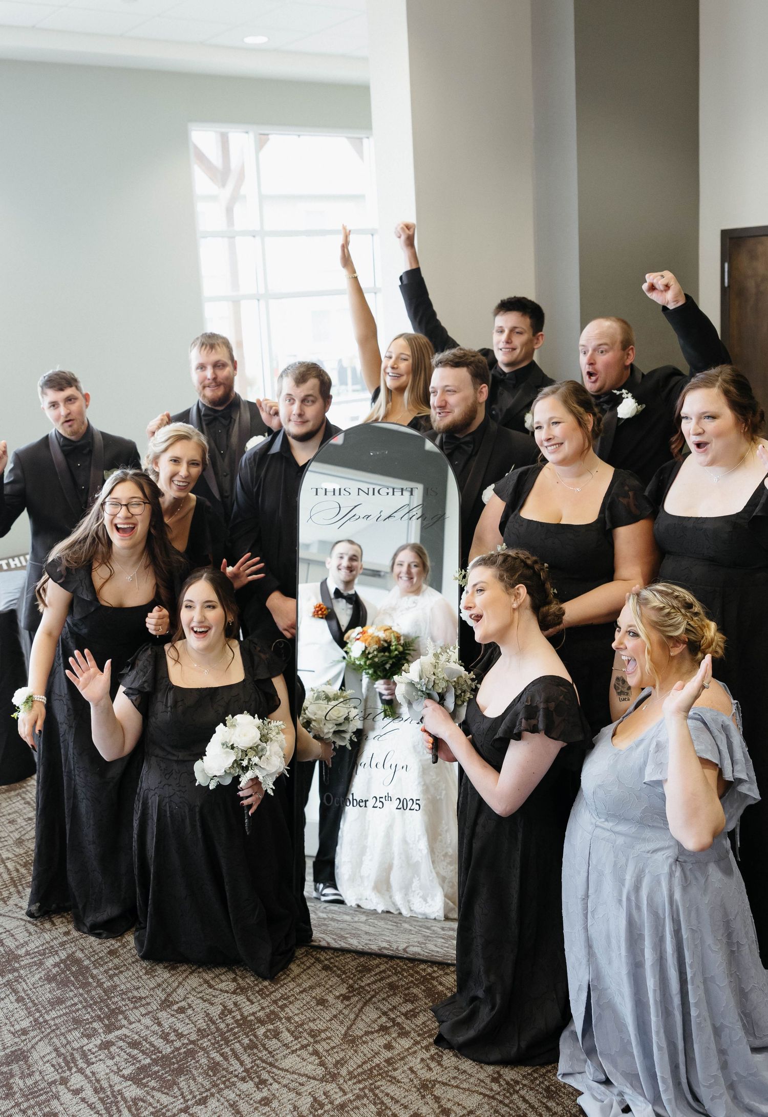 Wedding party members in black and gray dresses celebrate joyfully in a bright indoor venue.