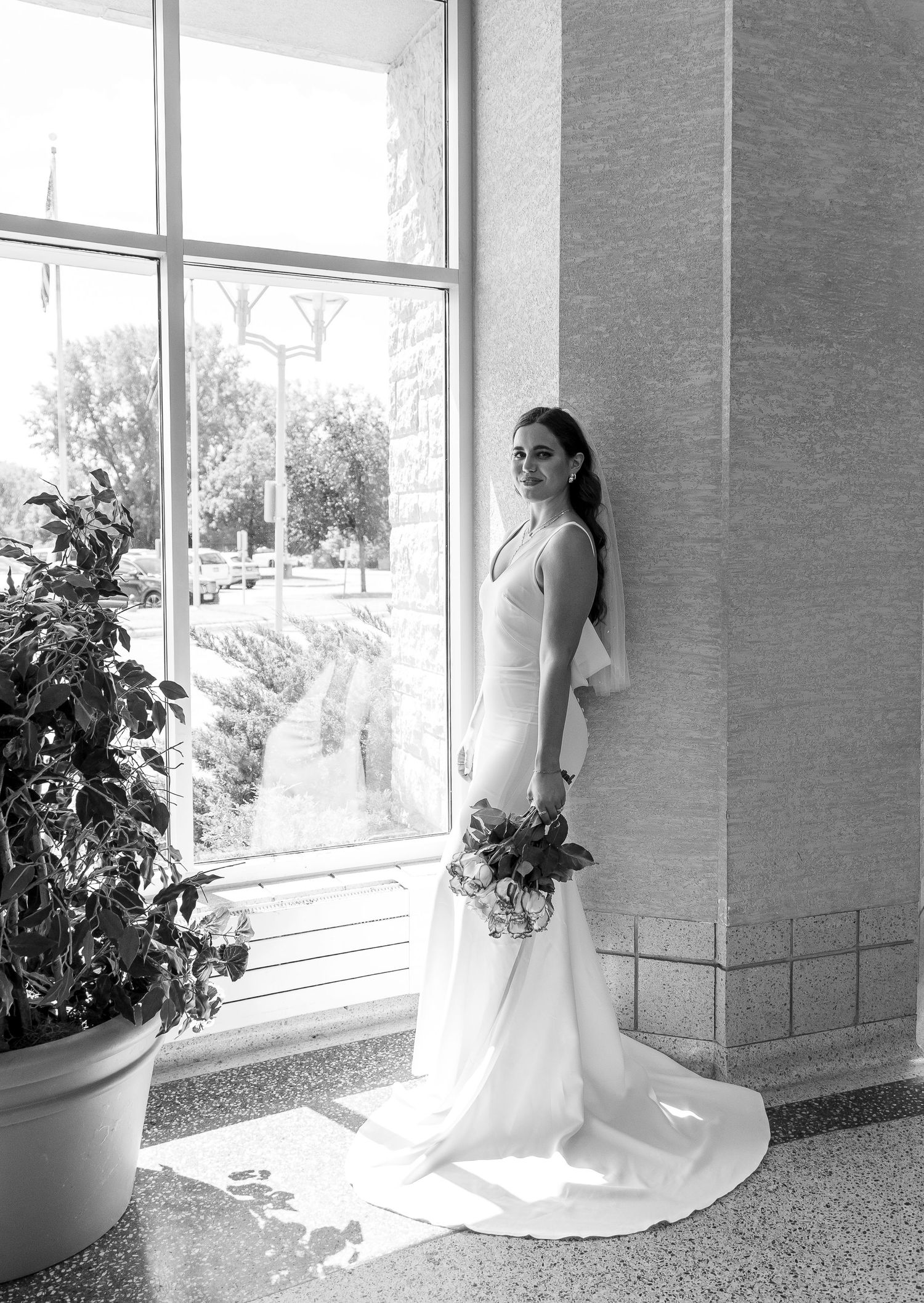 Bride in flowing white dress poses near large windows in a modern interior space.