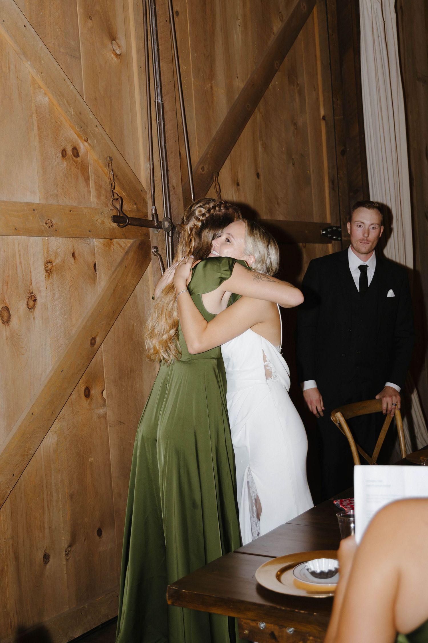 Two people embrace at a rustic barn wedding reception while another guest looks on.