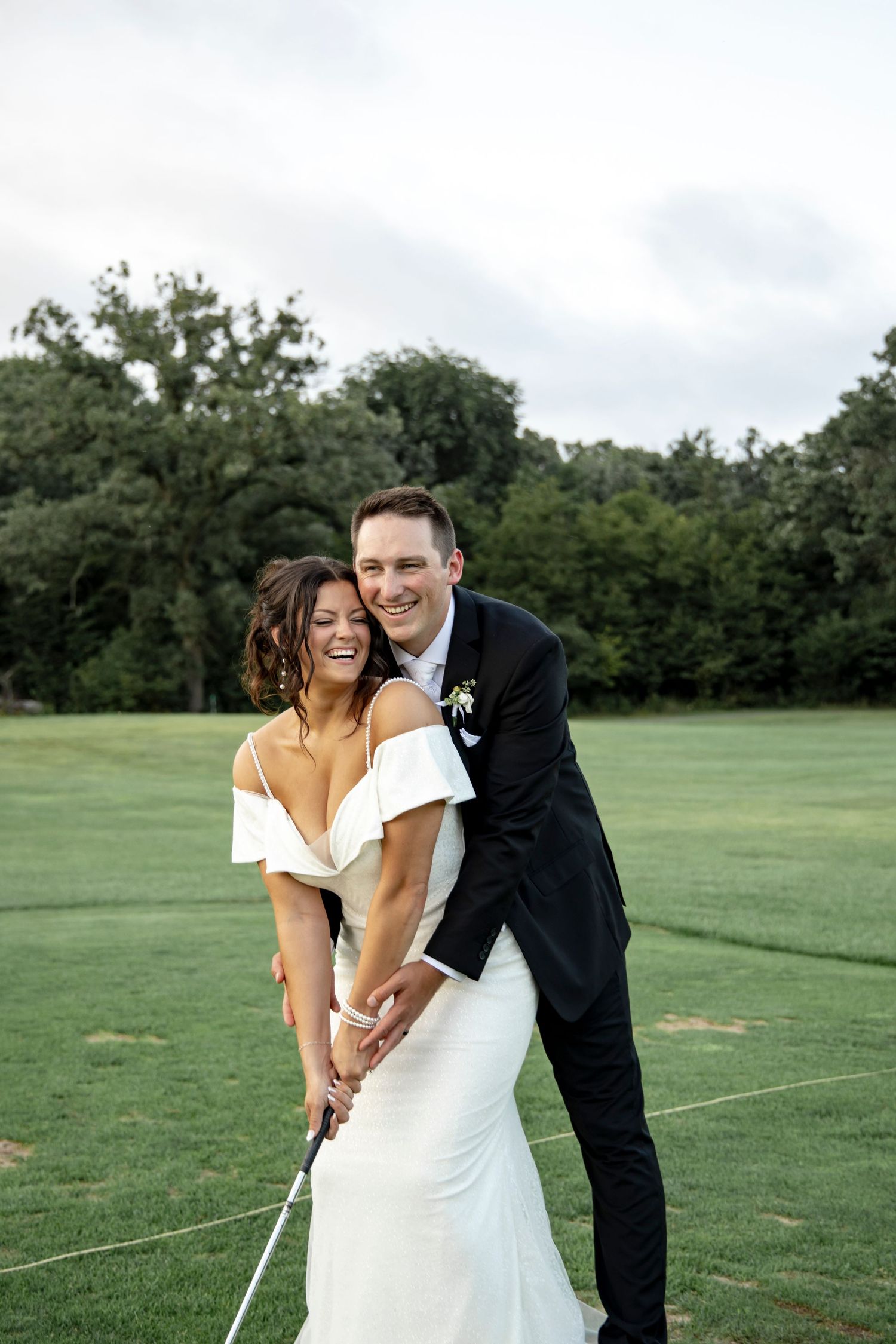 A playful wedding couple practices their golf swing together on a green lawn.