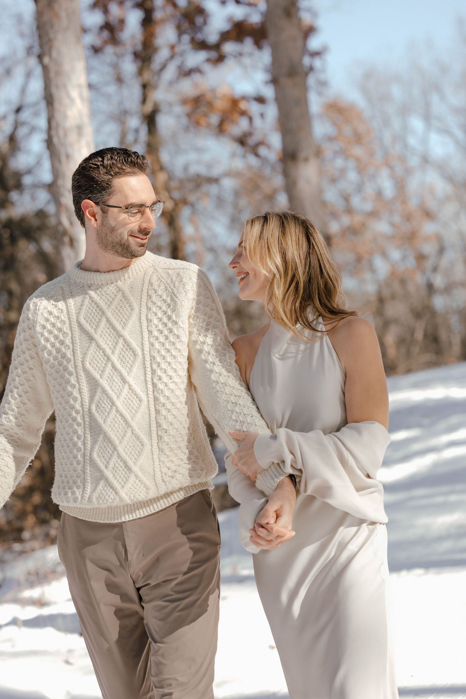 A couple walking together in winter, wearing coordinated cream and beige outfits against a snowy backdrop.