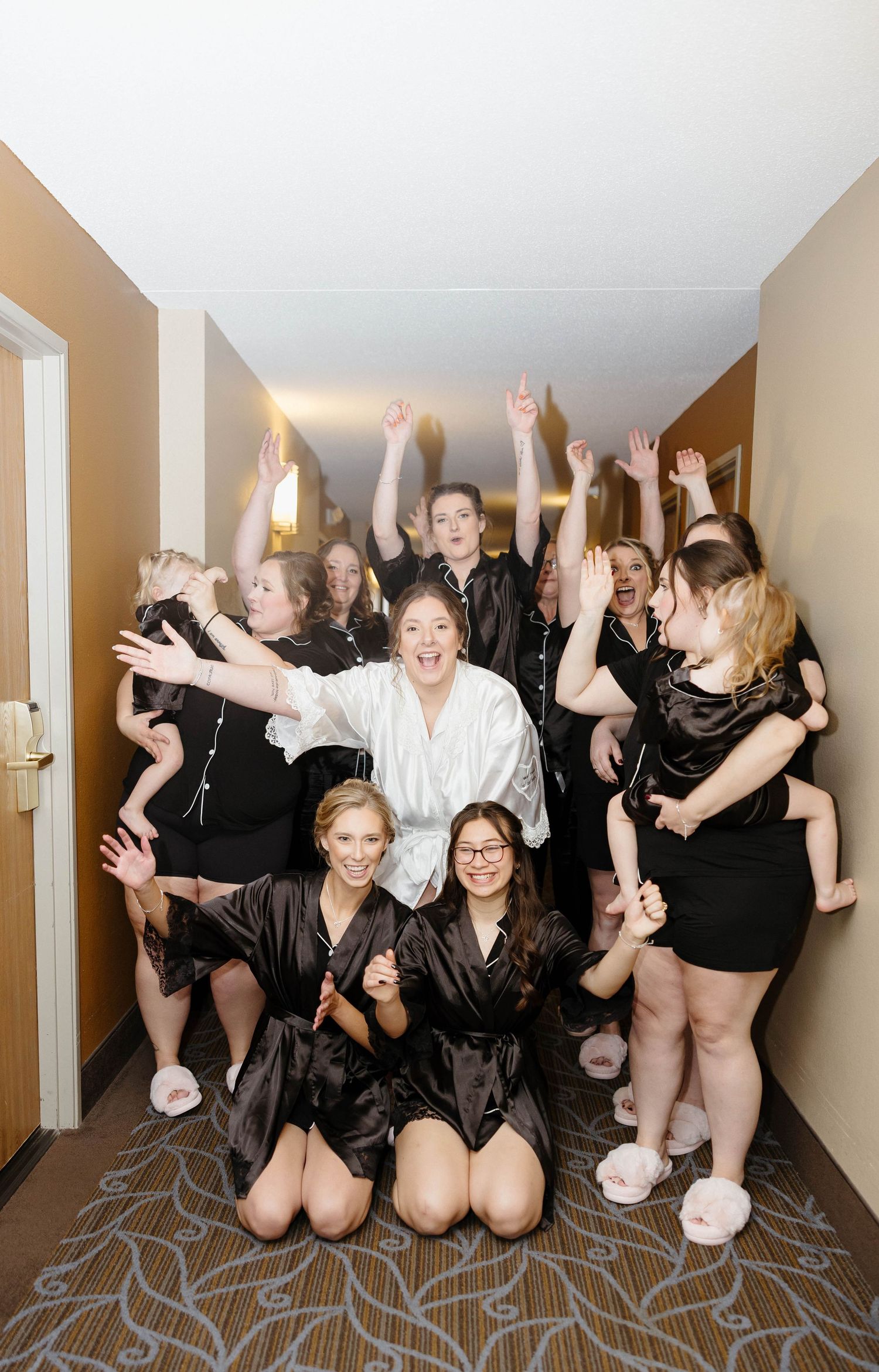 Bridesmaids in black robes pose energetically on hotel room floor.
