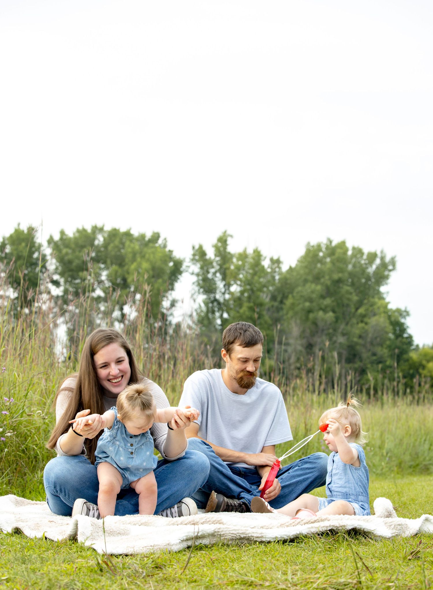 Family lying together on a white blanket in a sunny meadow with trees in the background.
