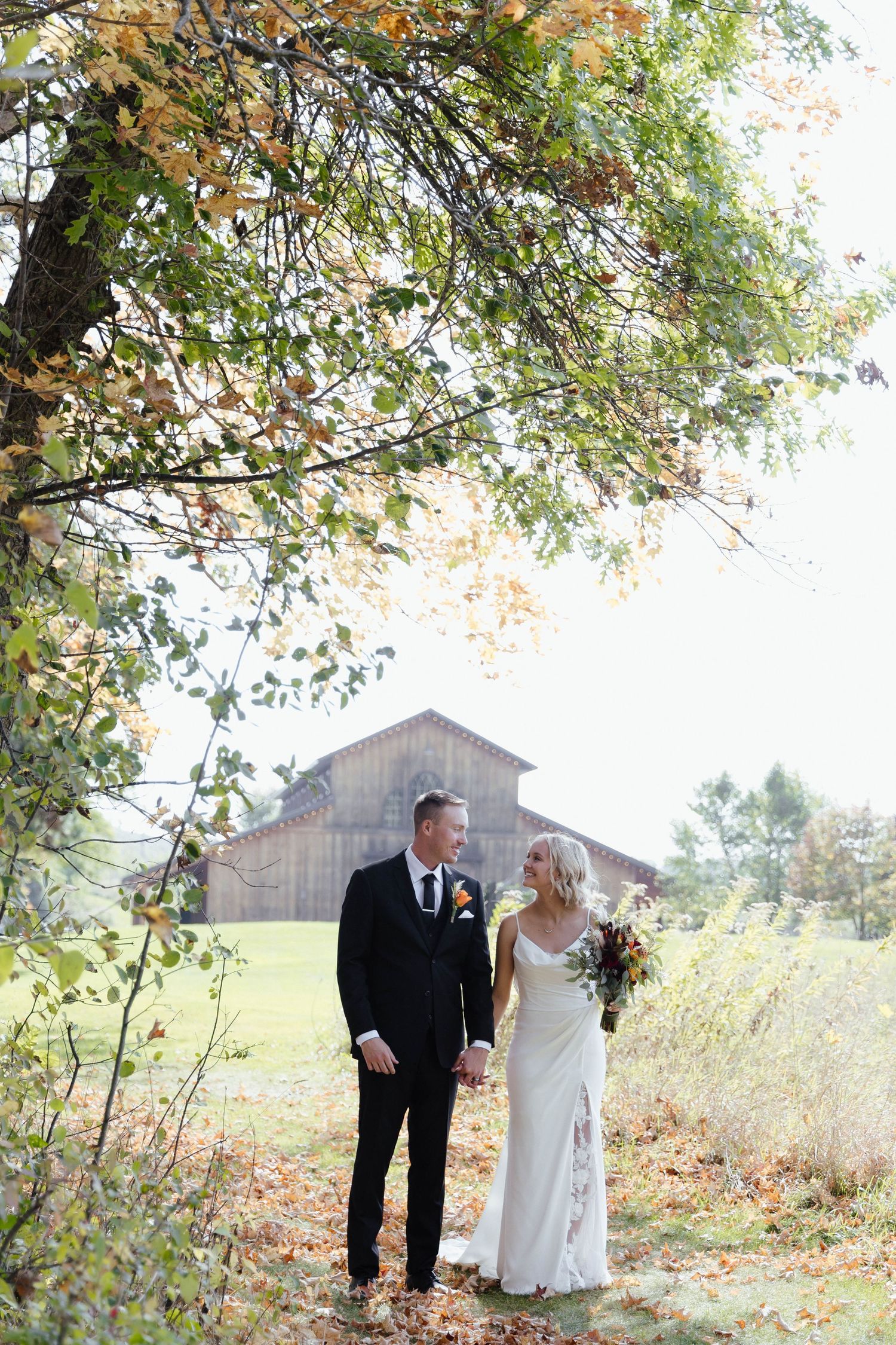 Newlyweds walking hand in hand through an autumn field with rustic barn in background.