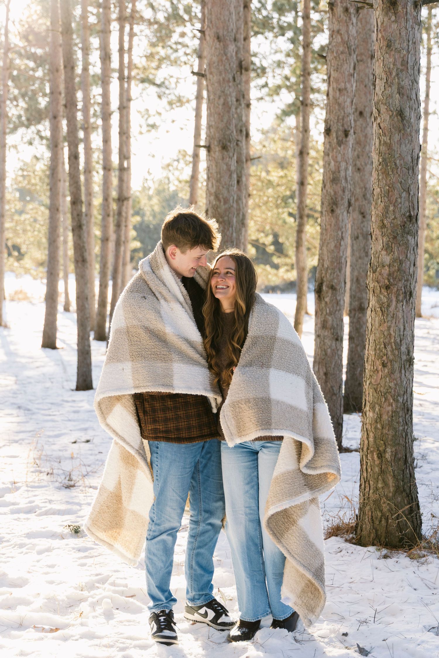 Couple wrapped in blanket sharing tender moment in snowy pine forest.