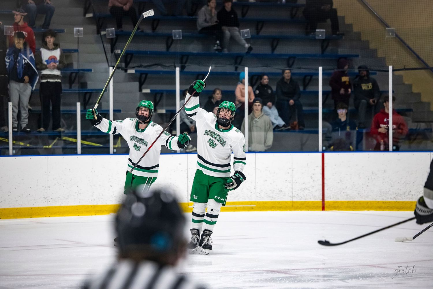 Ice hockey players in green and white uniforms chase the puck along the boards.