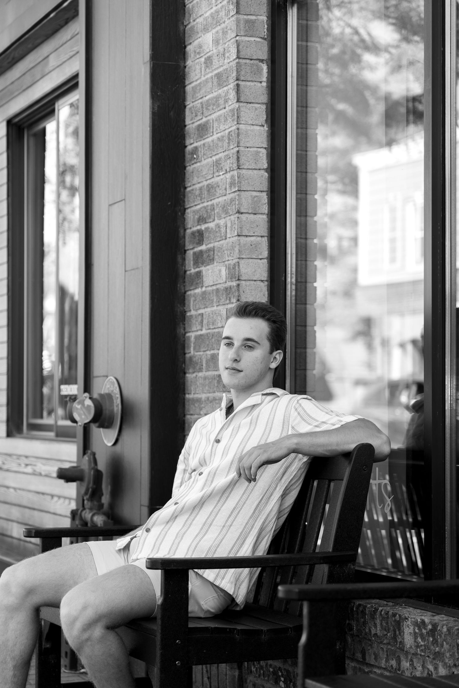 Person relaxes in wooden chair on brick building porch in black and white photo.