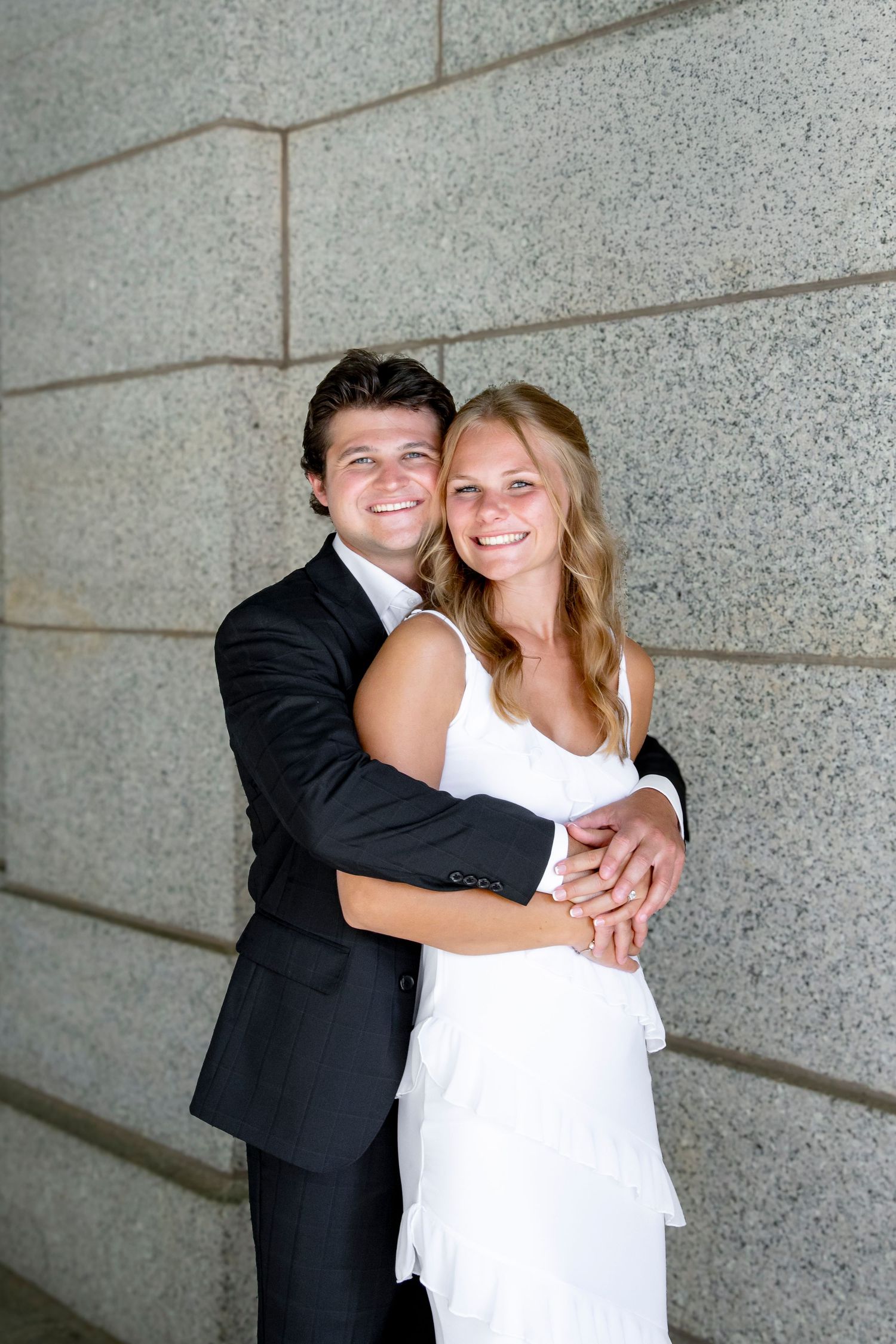 A couple in formal attire embracing against a textured stone wall background.