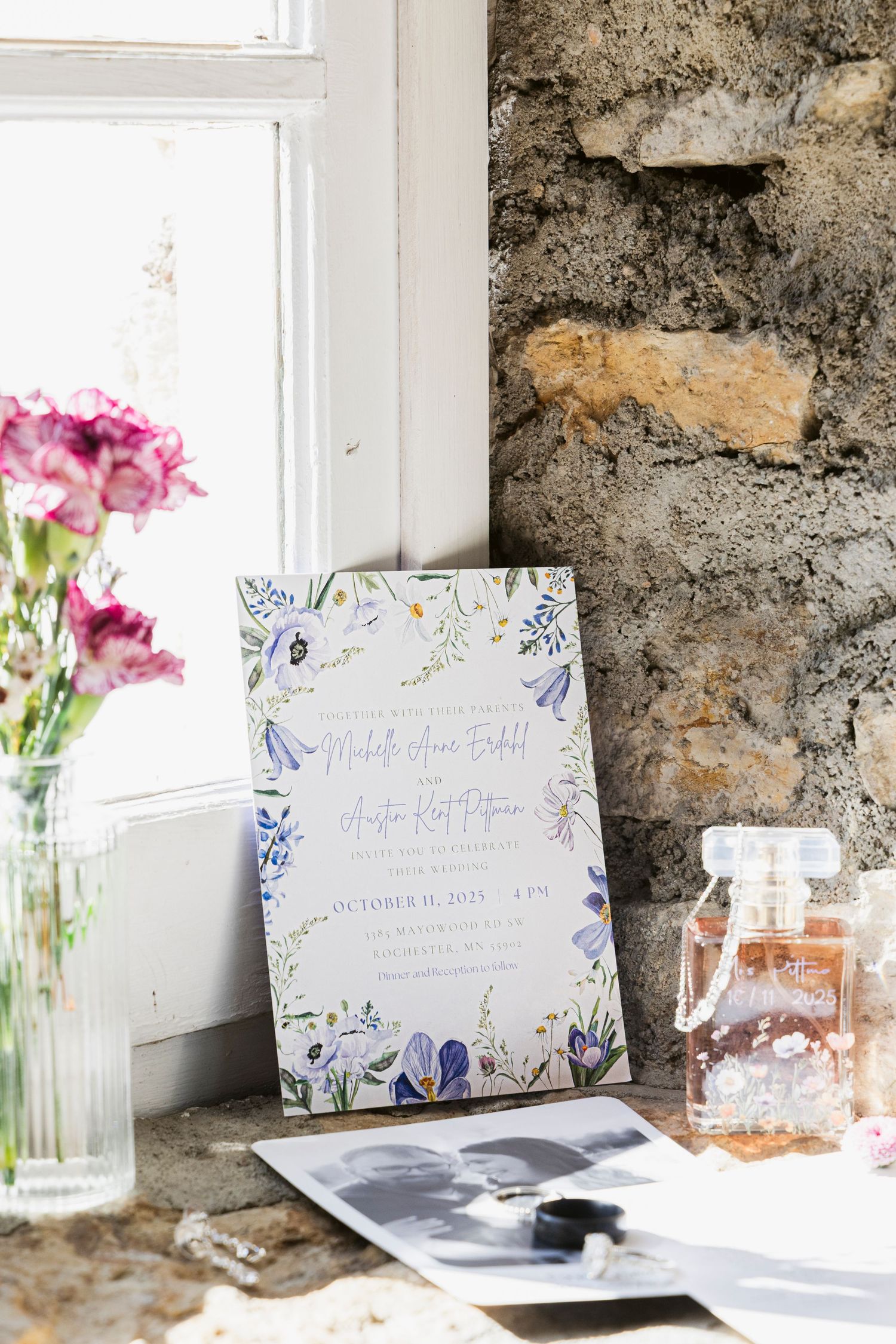 Wedding invitations and floral decorations arranged on a rustic windowsill with stone wall backdrop.