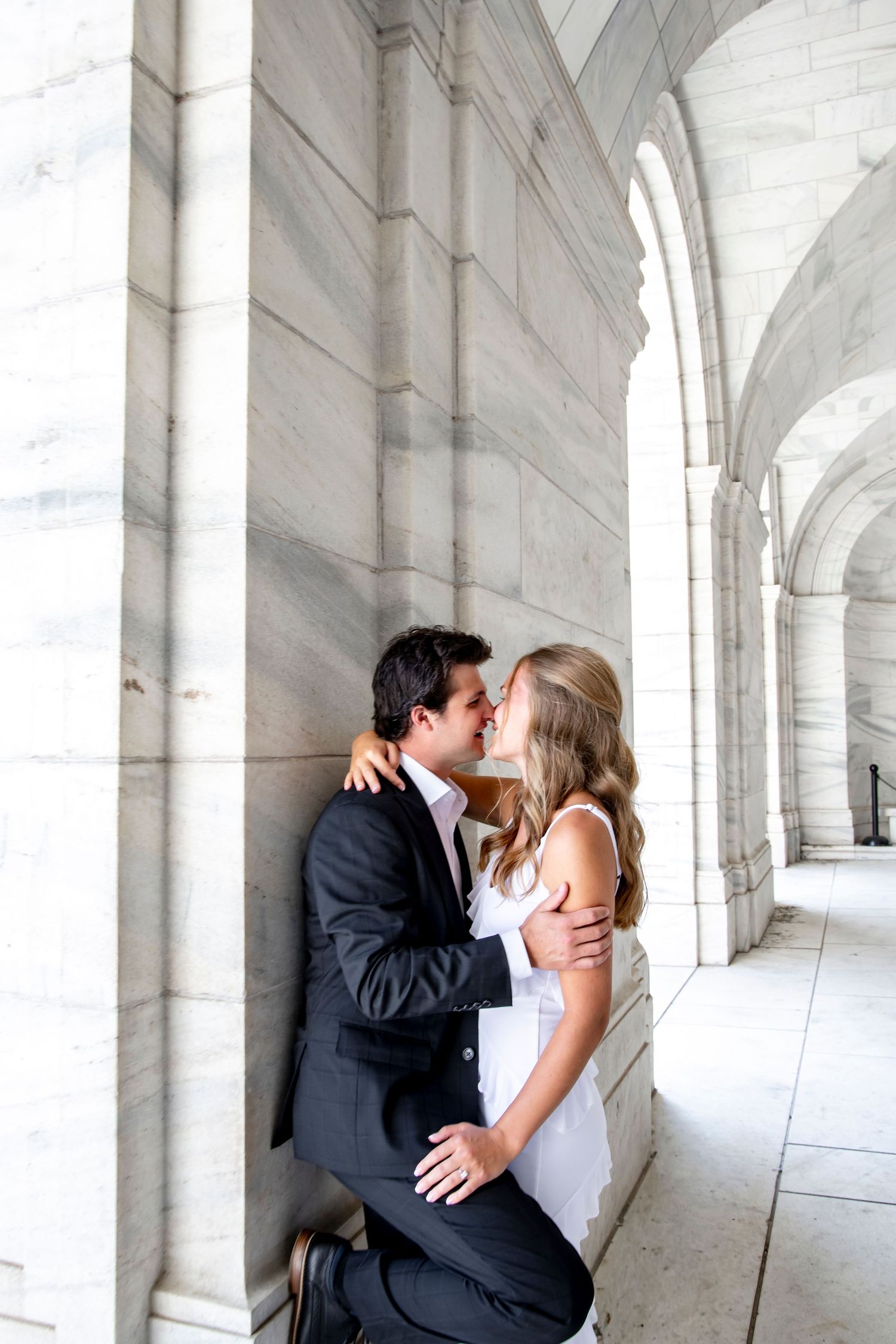 Romantic couple sharing intimate kisses in an elegant white marble colonnade hallway.