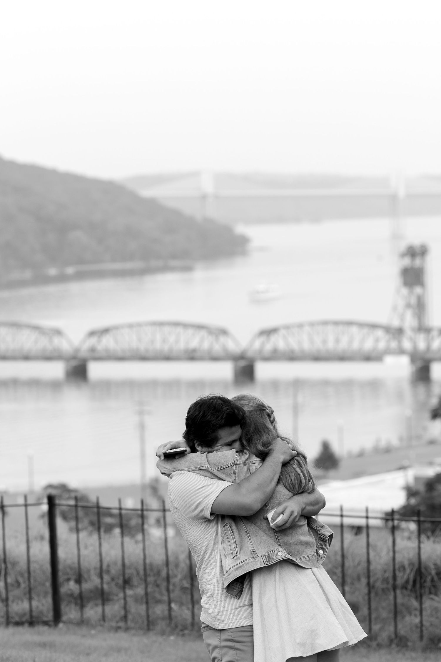 A couple embracing with a scenic river and bridge landscape in the background.