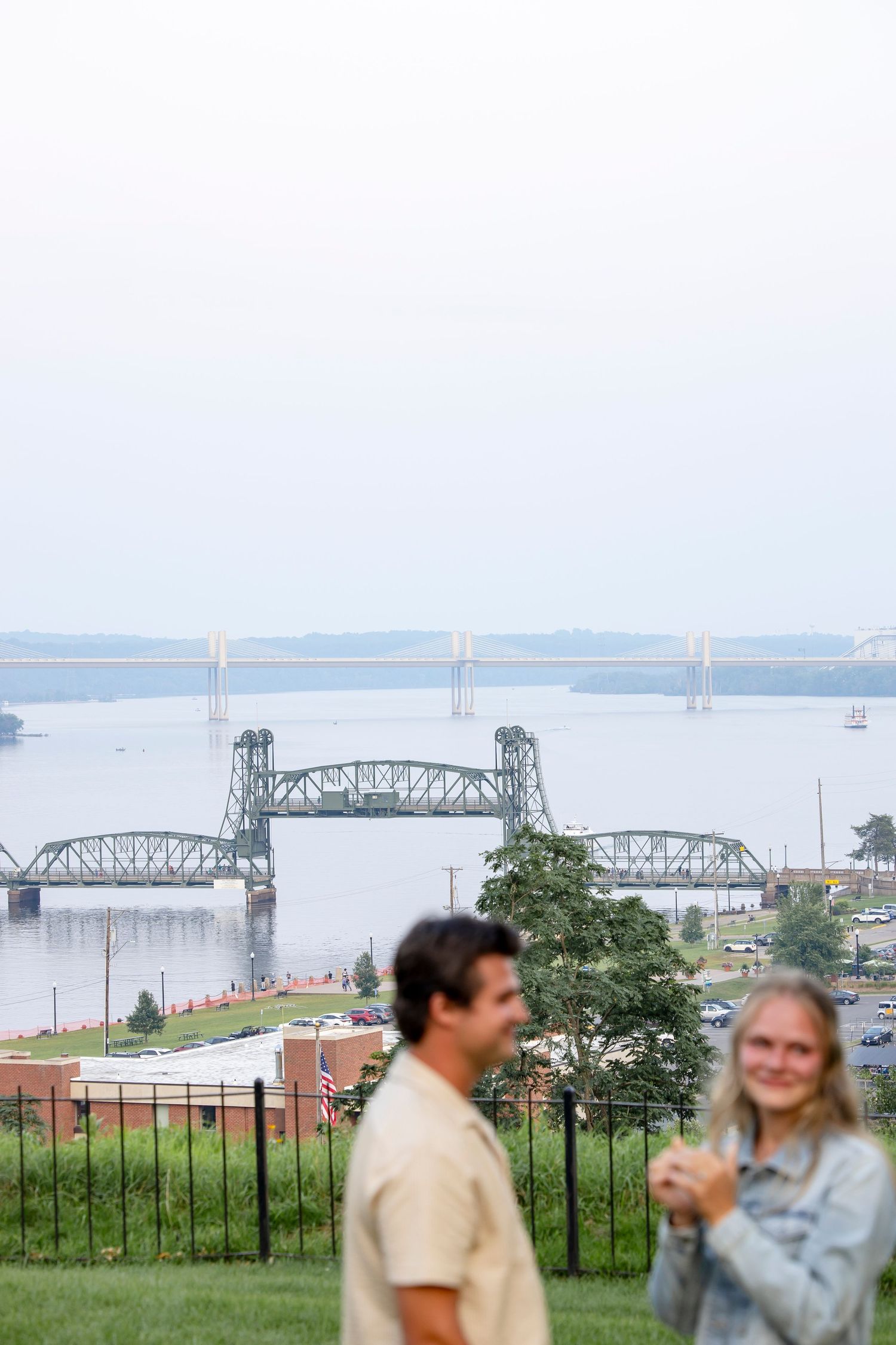 A couple enjoys a picnic with scenic views of a bridge and river in the distance.