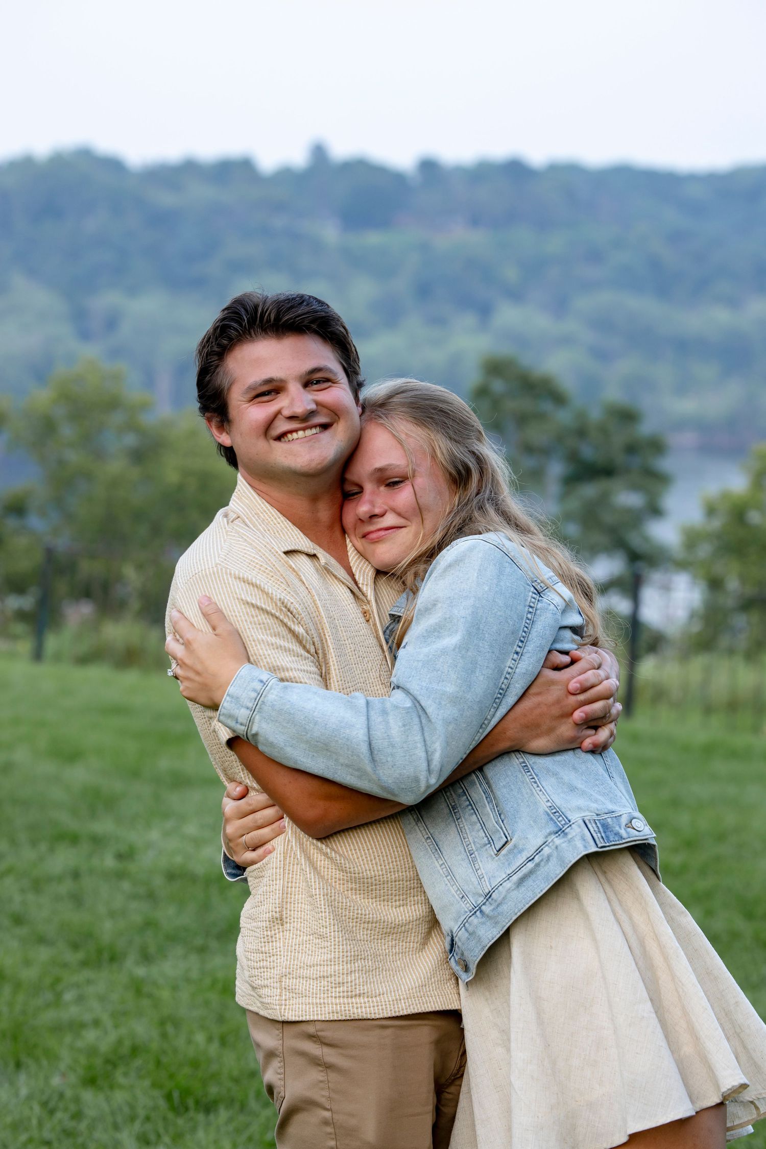 A romantic couple embraces in a grassy field with rolling hills in the background.