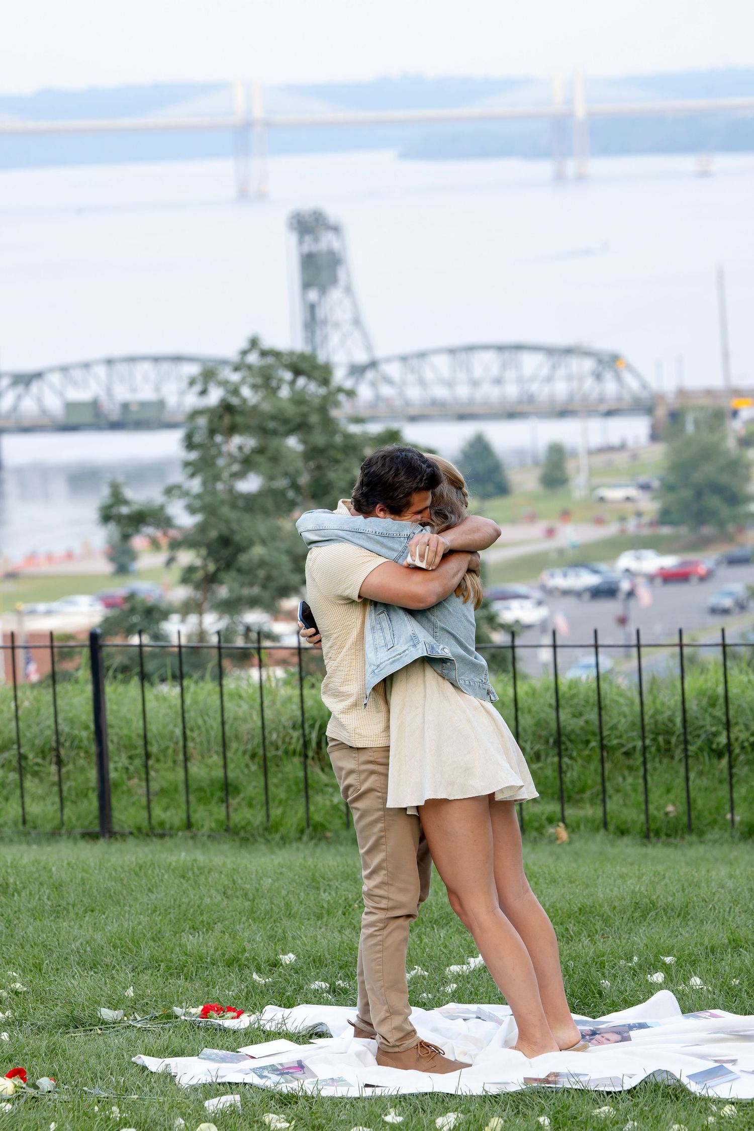 A couple embracing on a blanket with a bridge and river visible in the background.