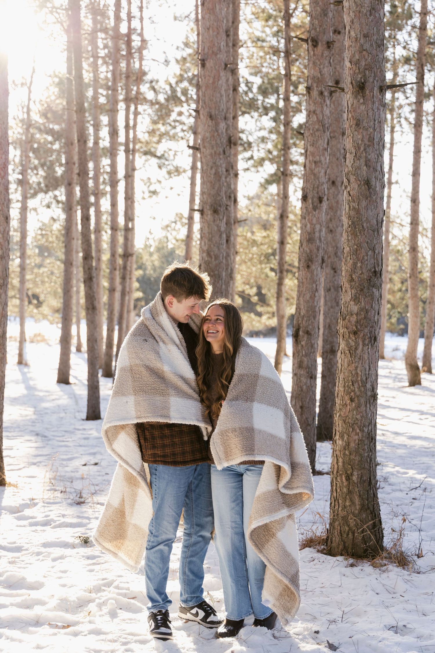 Couple wrapped in blanket shares intimate moment in snowy pine forest.