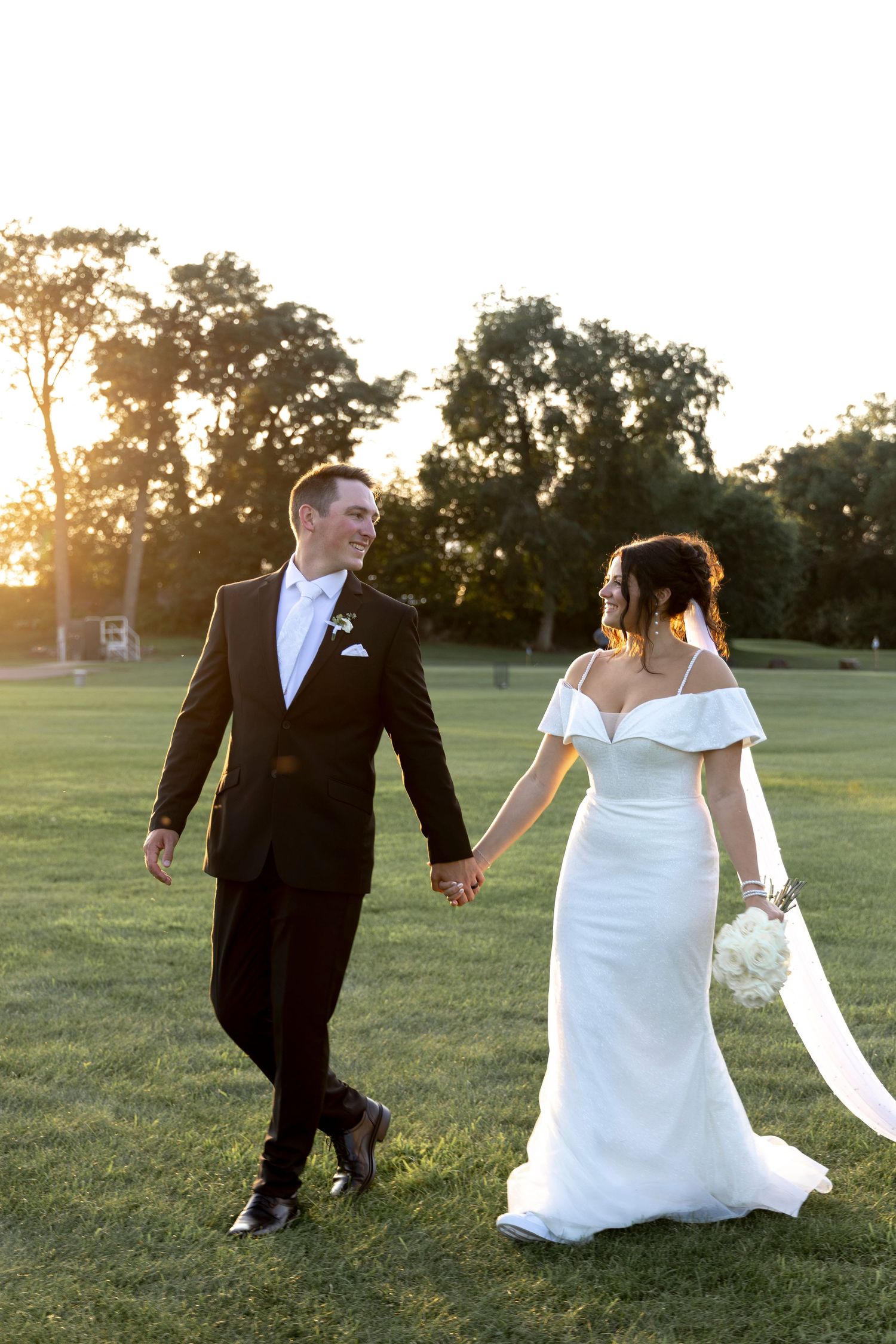 Newlyweds walking together on grassy field during sunset.