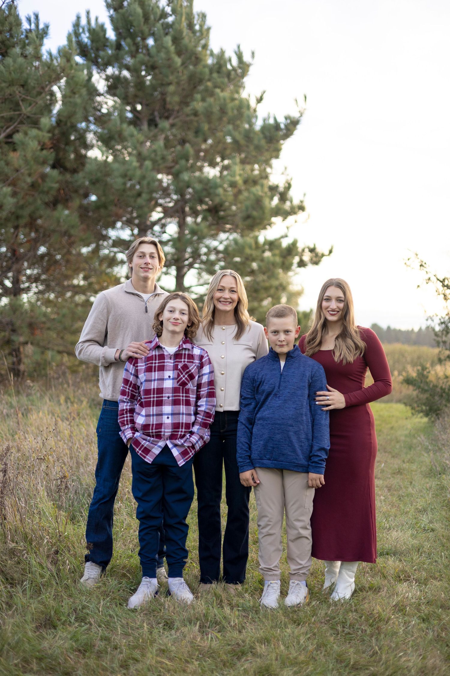 Family members stand together in casual outfits for outdoor group portrait with pine trees.